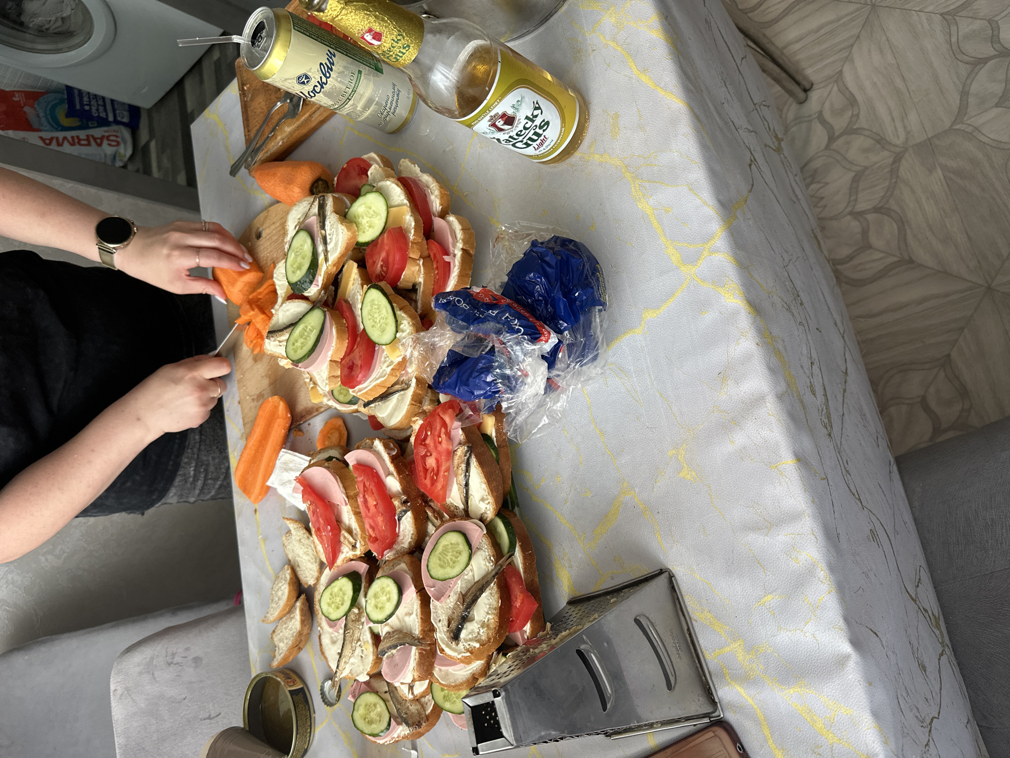 A person, possibly female given the jewelry on their left wrist, is preparing food on a table covered with a marble-patterned tablecloth. The person is wearing a dark t-shirt and is in the process of slicing a carrot on a wooden cutting board. Several prepared sandwiches, made with white bread, are stacked on the table. These sandwiches appear to be topped with cream cheese, cucumber slices, tomato slices, ham, and small fish, possibly sardines or sprats. A can of beer with a straw inserted is visible to the right of the sandwiches. A glass bottle of beer, also to the right, has a yellow label. A metal grater and an open can of what appears to be oil or broth are on the left side of the table. The background shows a washing machine, a bag of detergent labeled "SARMA," and a gray chair. The floor has a patterned tile design. The scene appears to be indoors, likely a kitchen or dining area, and is not in the city of Вознесенка, Russia.