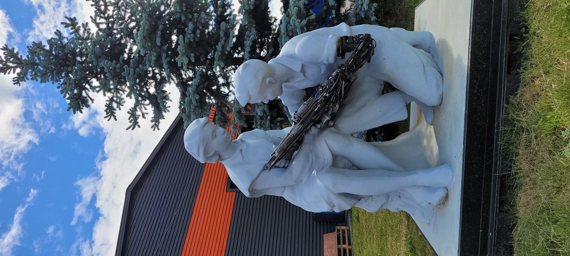 The image shows a marble sculpture depicting two figures, one cradling the other, with a metallic object resembling a firearm across their arms. The sculpture is positioned outdoors, set against a backdrop of a pine tree, a building with a gray exterior and a vertical orange stripe, and a blue sky with white clouds. The location is Unknown, Unknown.