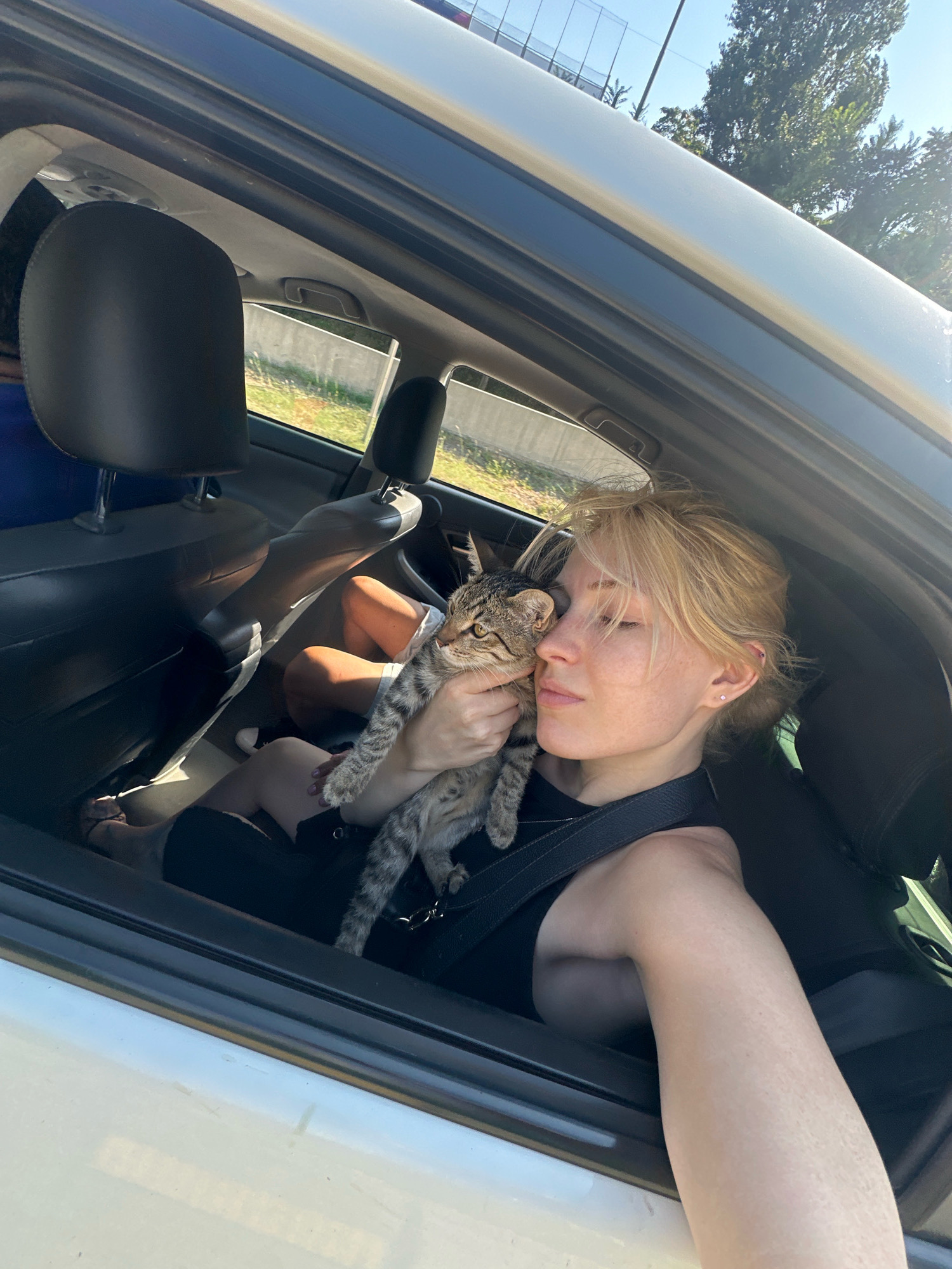 A young woman with blonde hair, wearing a black top and a seatbelt, is holding a tabby cat in her arms inside a car. The cat is being held close to the woman's face, and both appear to be looking forward. The woman's eyes are closed, and she has freckles on her face. Another person is visible in the back seat, partially obscured by the woman. The interior of the car, including headrests and seats, is visible, along with a portion of the car's exterior and the outdoor environment through the windows. Sunlight streams into the car, illuminating the subjects. The image appears to be taken from inside the car, looking towards the driver's side and the front passenger area. The exterior shows trees and a fence, suggesting a semi-urban or suburban setting.