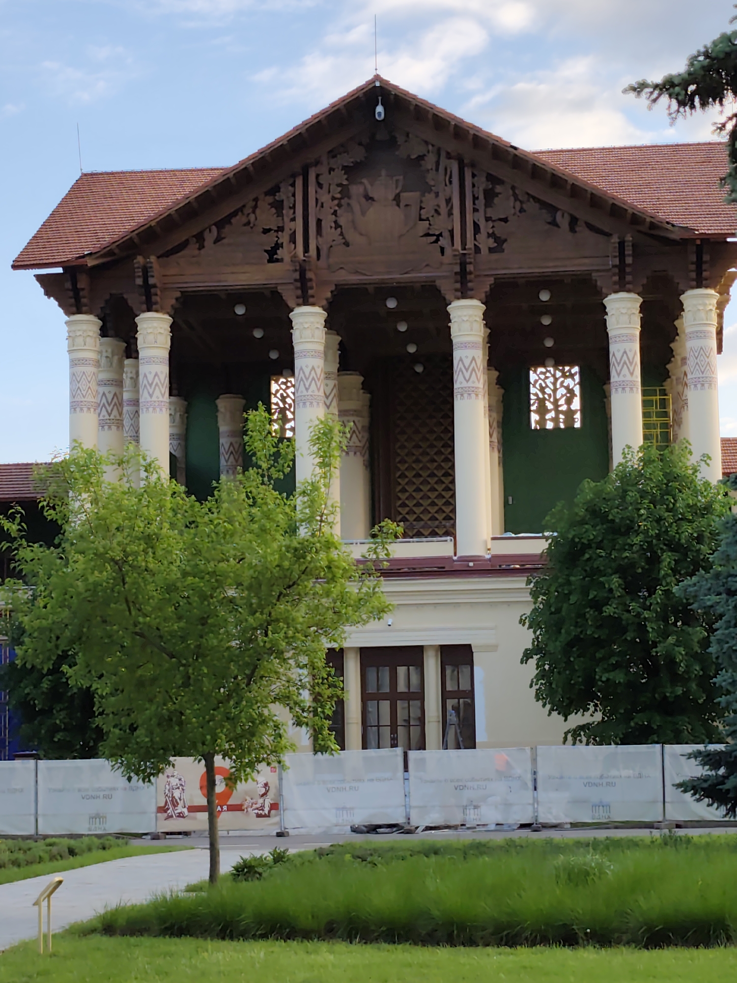 The image features a building in Moscow, Russia. The building has a light-colored facade, with a second story supported by white pillars adorned with patterned decorations. The top of the building is a dark wood with intricate carvings. The roof is a reddish-brown tile. In front of the building is a barrier with Russian writing on it. The foreground includes trees and grass. The sky is partially visible and appears to be a mix of blue and white.