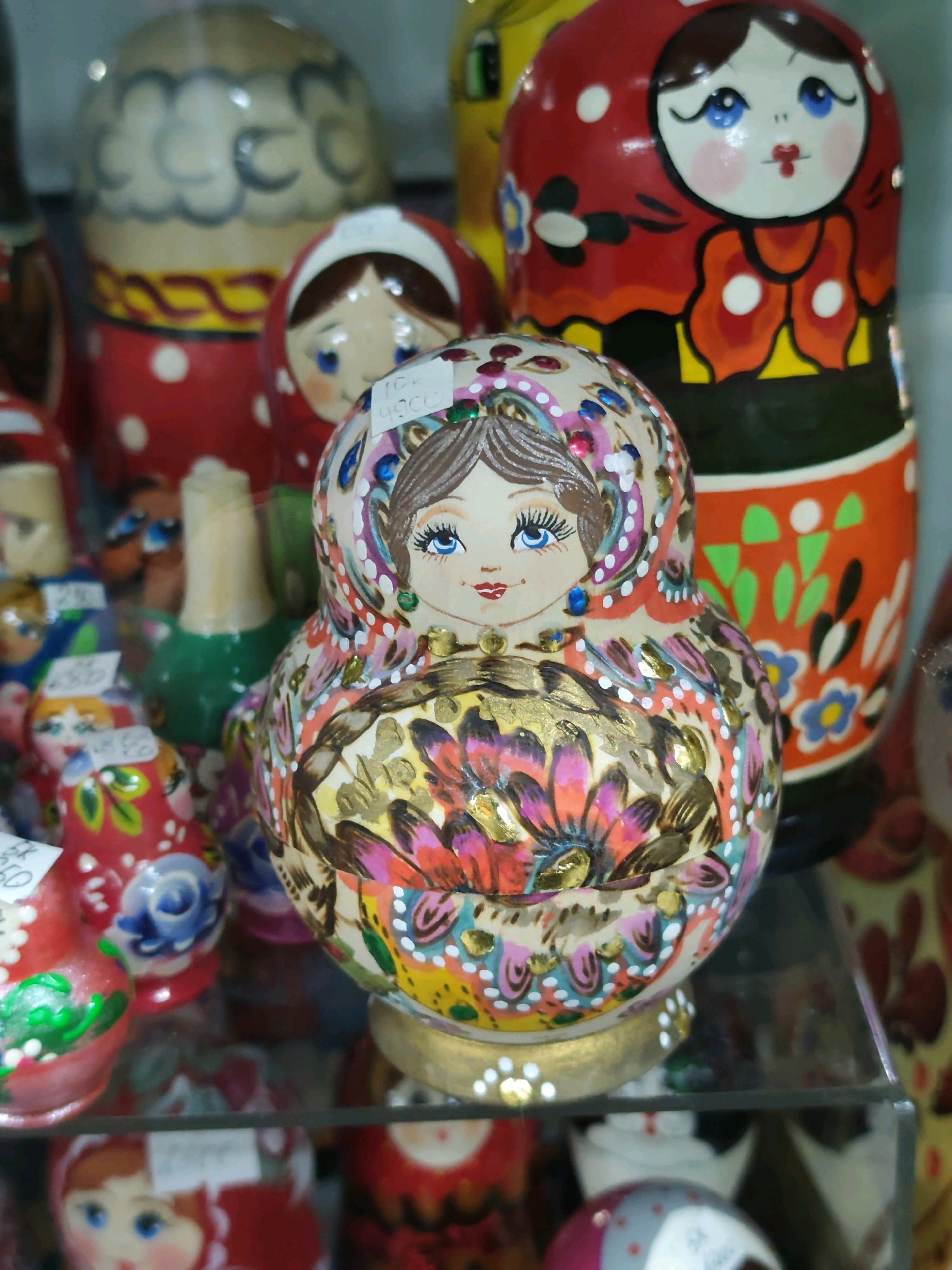 The image shows a close-up of several Matryoshka dolls displayed on a shelf, likely in a souvenir shop in Listvyanka, Russia. The dolls are intricately painted with traditional Russian designs. The focal point is a large doll in the foreground, adorned with vibrant floral patterns in shades of pink, orange, yellow, and blue, accented with gold paint. It has a delicately painted face with blue eyes and red lips. Behind it, another doll with a red headscarf and a blue-eyed face is visible. To the left and in the background, other dolls with varying designs and colors are partially visible. The dolls are arranged on a reflective surface, possibly a glass shelf, which adds depth to the scene. The lighting appears to be artificial, highlighting the colors and details of the dolls.