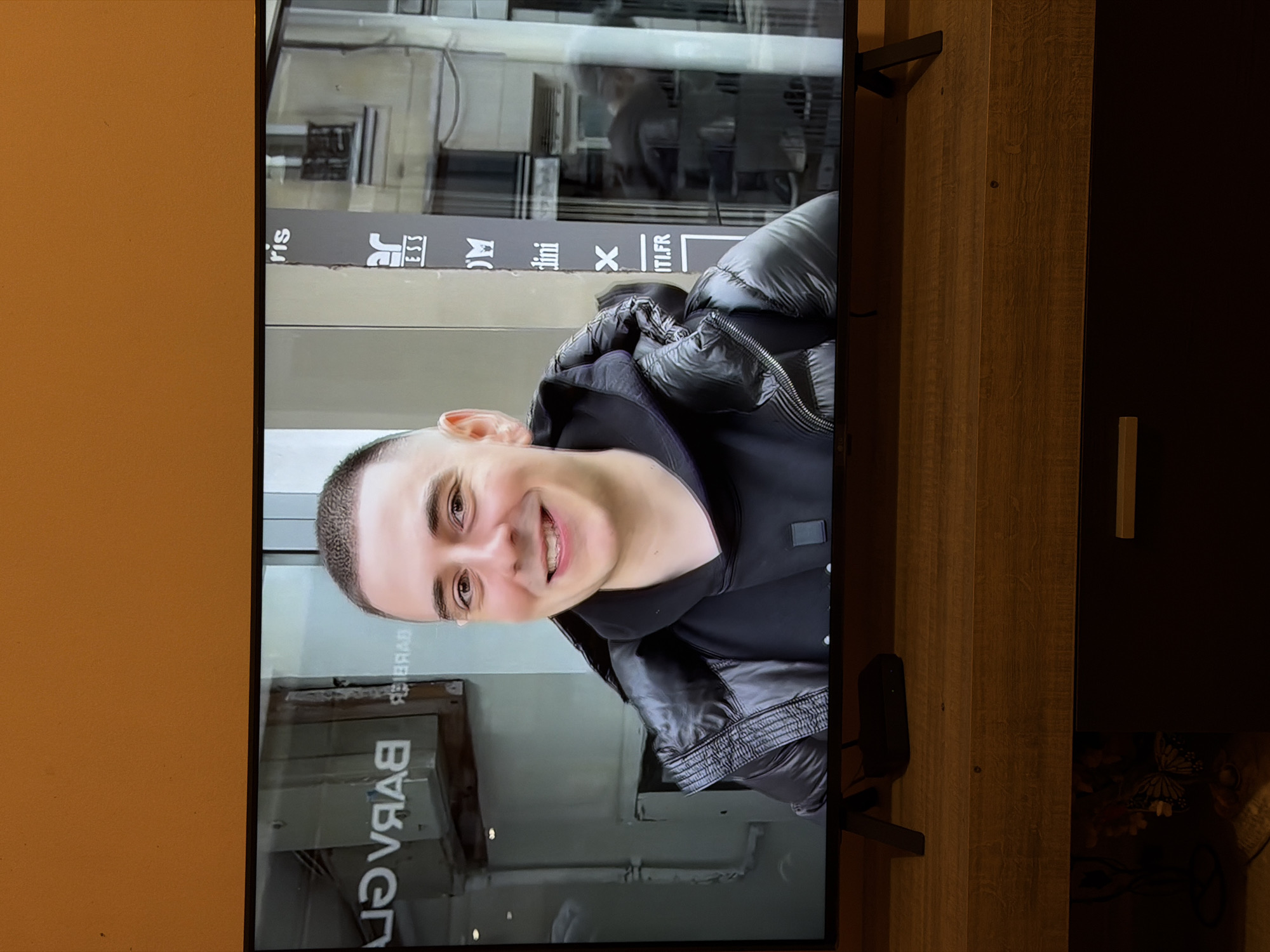 A light-skinned, smiling person with short hair is displayed on a flat-screen television. The person is wearing a black hooded top and a black puffer jacket. The television is positioned on a wooden TV stand, located in front of a dark cabinet with a light-colored handle. The reflected image on the TV screen shows a storefront with the word "BARVER" backwards, suggesting it is a reflection. The text above this is "ris Jerri martini X ITI.FR", indicative of a commercial establishment. The buildings in the background are typically found in an urban setting. The location appears to be a barber shop, potentially located in Sofia, Bulgaria. The environment seems to be indoors, possibly a living room. The person appears to be interacting with the camera by smiling.