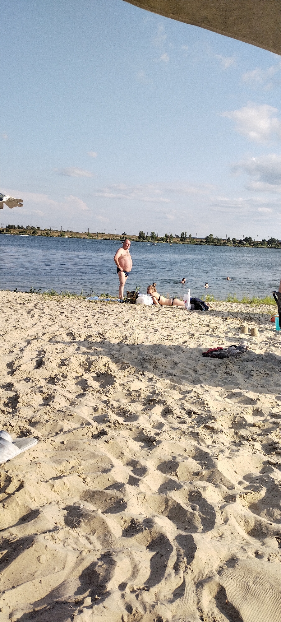 The image depicts a sunny day at a sandy beach beside a body of water, likely a river or lake, in Bezliudivka, Ukraine. The foreground is dominated by textured sand with shadows cast from an unseen object overhead, possibly an umbrella or canopy. In the middle ground, a man in dark swim trunks stands near the water's edge. To his right, a woman in a bikini is lying on her stomach on the sand. Several other people are visible in the water, swimming or wading. The background features a tree line along the opposite shore and a clear blue sky with scattered clouds. There are also some personal items scattered on the sand, including bags and footwear.