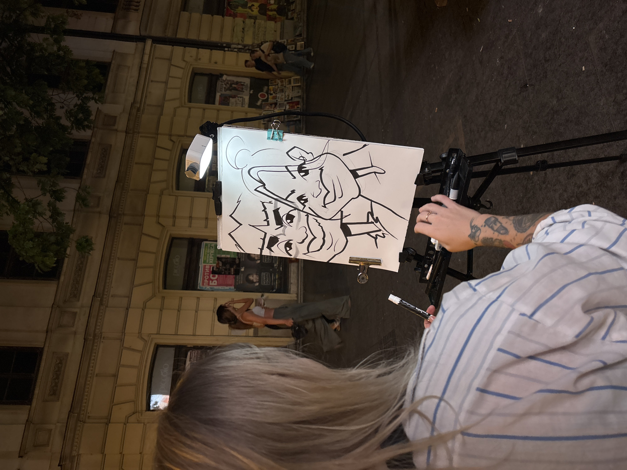 In Belgrade, Serbia, a blonde woman with tattoos on her arm is drawing a caricature on a sidewalk. The drawing depicts two people with exaggerated features, one with a large head and the other with a prominent chin. The woman is holding a marker and appears to be mid-drawing. She is wearing a blue and white striped shirt. The drawing is on an easel, illuminated by a small lamp. In the background, there is a building with shops, and other people are walking by. One shop has a sign that says "piccolo" in English. The scene is at night, and the streetlights are on.