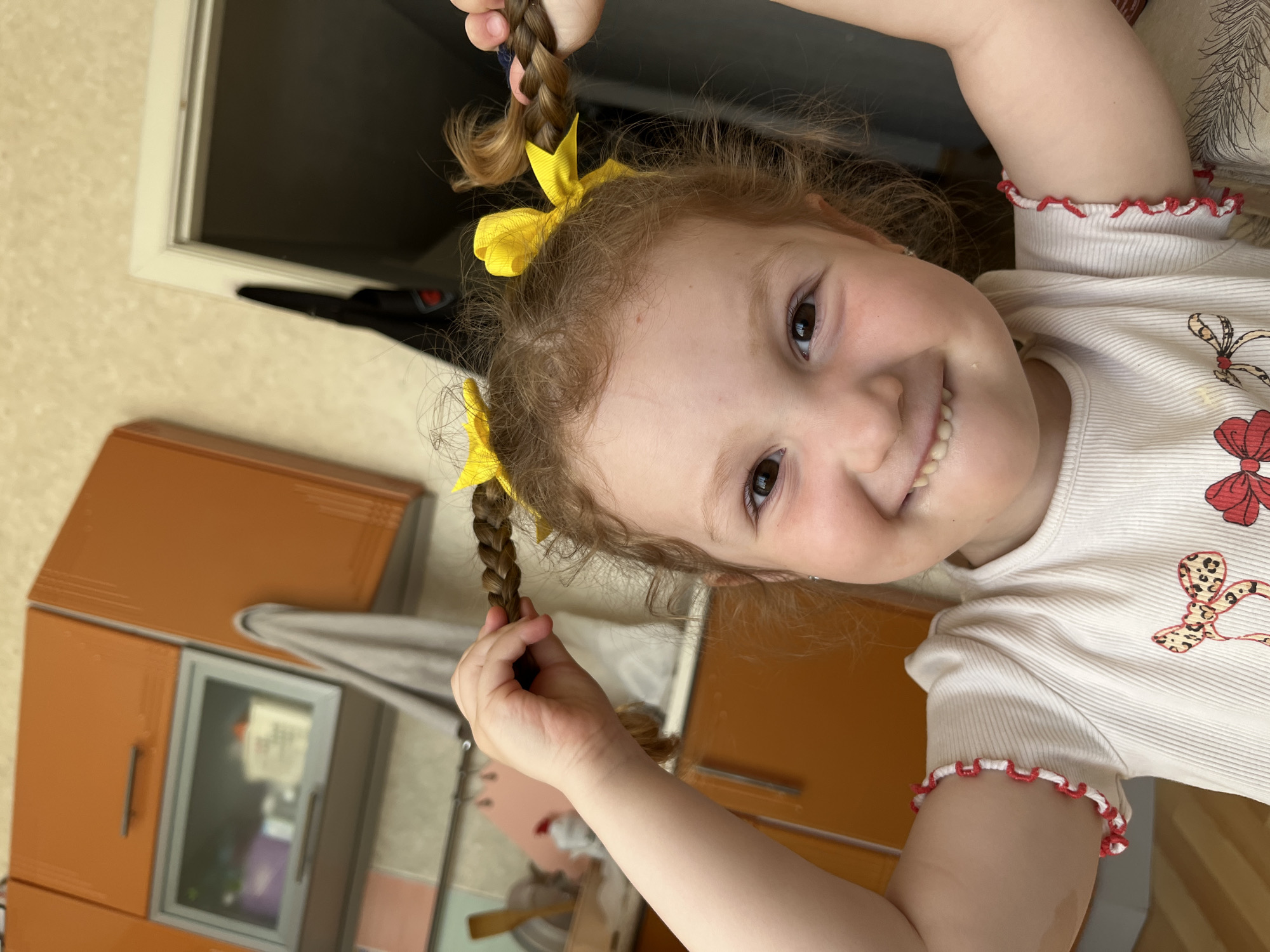 The image captures a young, light-skinned child, approximately 4-7 years old, with reddish-brown, curly hair styled into two braided pigtails, each secured with a yellow bow. She faces the camera directly, displaying a wide, open-mouthed smile that reveals both upper and lower teeth, including what appear to be developing permanent central incisors. Her cheeks are rosy, and her eyes are dark brown. The child wears a light-colored, possibly cream or off-white, ribbed short-sleeved top featuring multiple red and brown bow patterns on the front. On her upper left arm, a red and white coiled or ruffled armband is visible. Both of her hands are raised, each holding one of her braided pigtails.
The background indicates an indoor, domestic setting, likely a kitchen or an area adjacent to it. To the left, orange wall-mounted cabinets with metallic handles are present. One cabinet has a transparent panel, revealing blurry contents such as a purple container and other light-colored objects. A grey towel hangs from a fixture on another orange cabinet to the right. Above the cabinets, the wall is a light, possibly beige or off-white, color. To the far right, a white door frame leads into a darker interior space, and a dark fabric object, potentially a garment, hangs on the wall nearby.