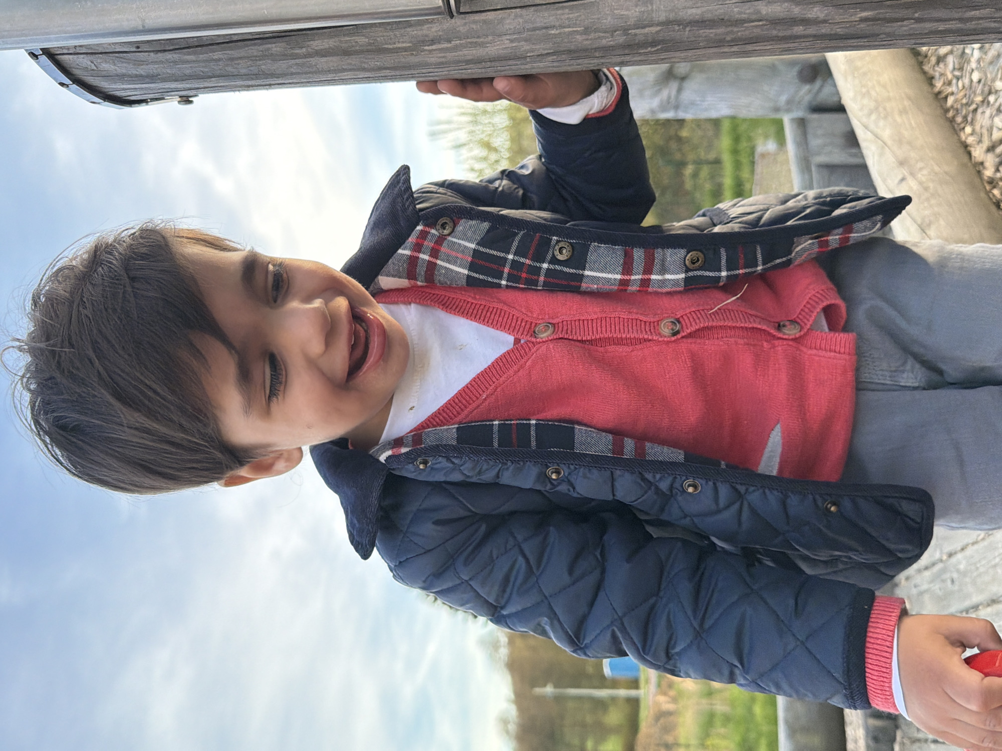 A young child is playing outdoors on a sunny day. He is holding onto a wooden structure, possibly part of a playground, and smiling at the camera. He is wearing a white shirt, a red cardigan, and a navy blue quilted jacket with a plaid lining visible at the collar. The environment appears to be a park or recreational area in Junglinster, Luxembourg, with green grass and trees in the background. The sky is partly cloudy.