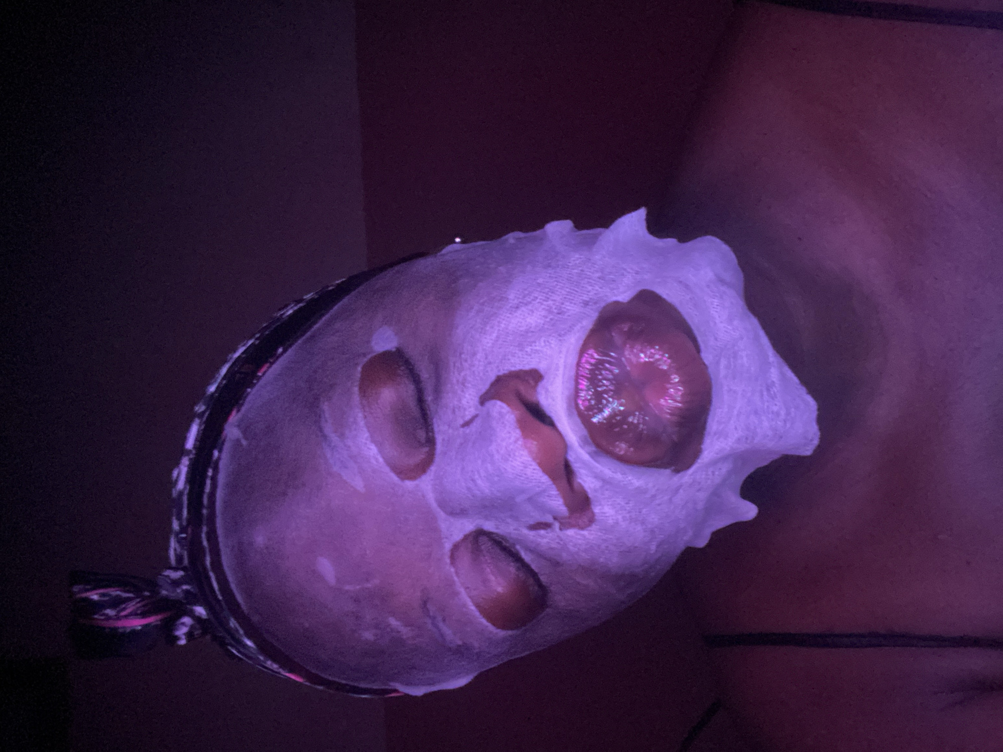 The image features a person, partially visible, wearing a white facial sheet mask in a dimly lit environment, bathed in strong purple light. The mask has cutouts for the eyes, nose, and mouth. Through the mask's left eye opening, a highly detailed, iridescent, multi-hued eye is visible, appearing artificial or part of an elaborate costume or prosthetic, rather than a natural eye. The skin visible through the mask cutouts and on the exposed upper body/shoulder area appears a reddish-brown tone, consistent with human skin under purple light. A dark band, possibly a hairband or a patterned headpiece, is visible on the left side of the person's head.
The scene suggests an indoor setting, likely at night or in a darkened room, given the artificial purple illumination. The activity is either a beauty treatment involving a face mask or participation in an event requiring unique artistic makeup or costuming, highlighted by the striking artificial eye. The dominant purple lighting creates a mysterious and artistic ambiance. No specific location cues for Maigizo, Nigeria, are visible within the image itself. No text is visible.