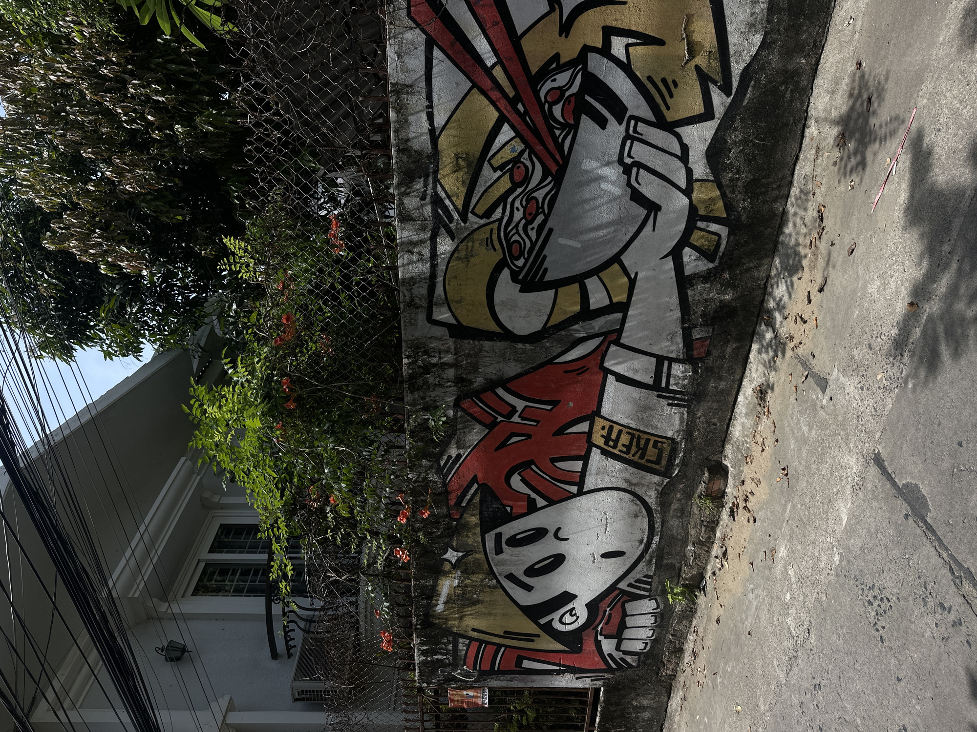 A graffiti-covered concrete wall runs along a sun-drenched street in Bac Nha Trang Ward, Vietnam. The graffiti features two cartoonish figures. The left figure has a large, white head with black oval eyes and a blank expression, wearing a red and white outfit. A tag reading "SKER" is visible on its chest. The right figure is mostly yellow and appears to be holding a bowl of noodles with red chopsticks. Orange trumpet flowers bloom above the wall, and lush green foliage is visible behind a chain-link fence. The street in front of the wall is asphalt, littered with dry leaves and shadows cast by overhead trees and power lines. A white building with a balcony and an air conditioning unit is visible behind the foliage on the left.