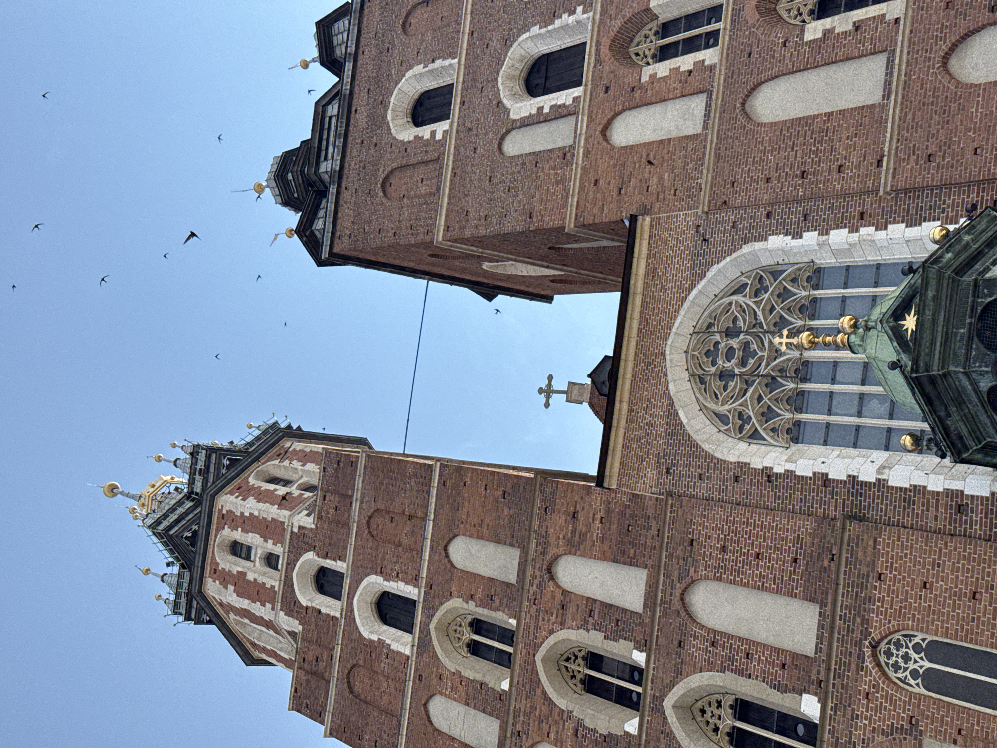 This image shows a low-angle view of the towers of St. Mary's Basilica in Krakow, Poland, against a clear blue sky. Several birds are visible flying in the sky above the basilica. The basilica is constructed of red brick and features Gothic architectural elements, including arched windows with intricate tracery and decorative spires. The main facade has a large rose window above a prominent arched doorway. A golden cross on a pedestal is visible on the rooftop section between the two towers. The lighting suggests it is daytime, with the sun illuminating the brickwork.