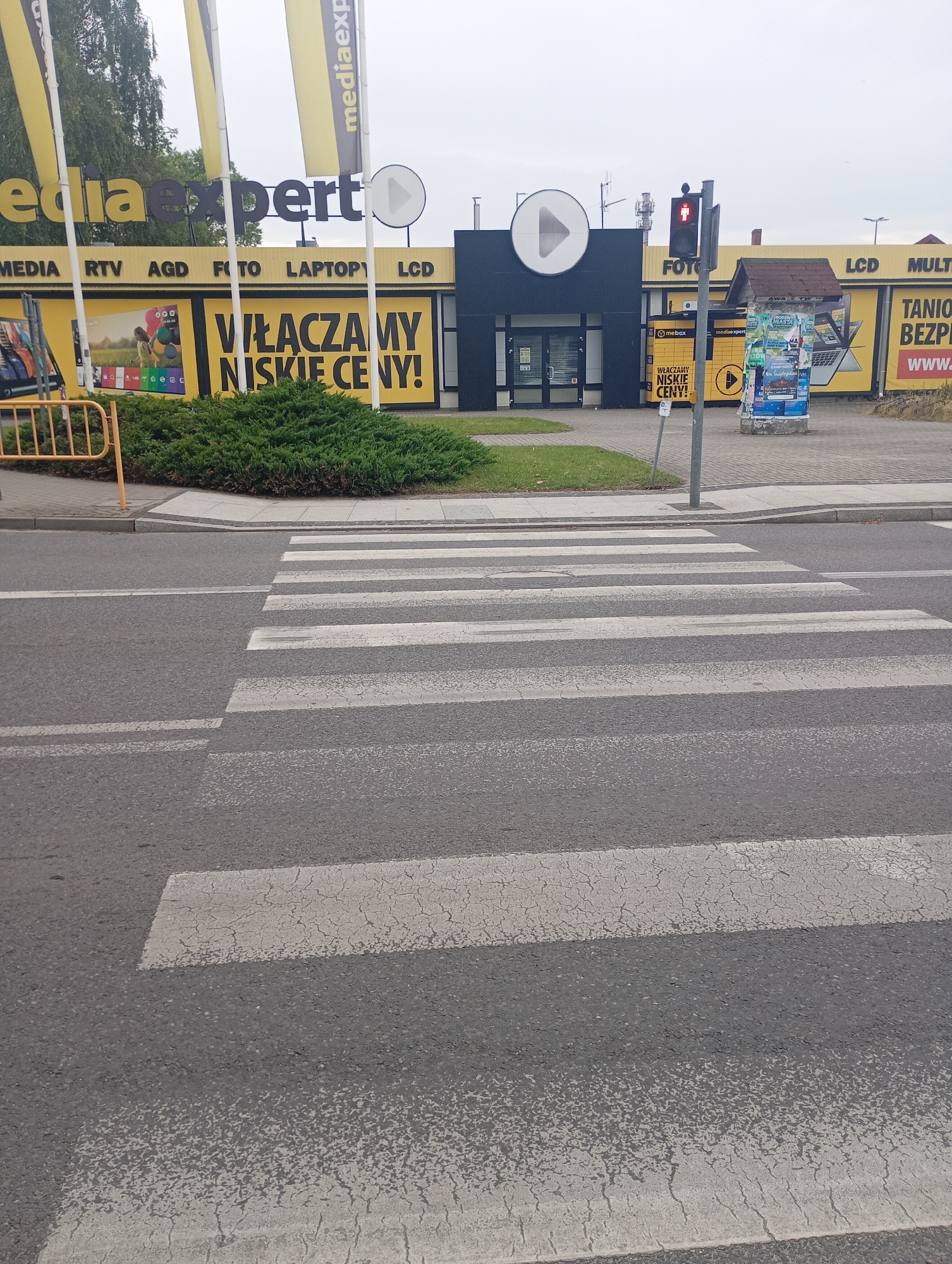 The image depicts a crosswalk in front of a Media Expert store in Międzyrzecz, Poland. The Media Expert storefront features large yellow banners with black lettering advertising various electronics categories such as "MEDIA", "RTV", "AGD", "FOTO", "LAPTOPY", and "LCD". A prominent slogan in large black letters reads "WŁĄCZAMY NISKIE CENY!" which translates to "WE TURN ON LOW PRICES!". A traffic light with a red pedestrian signal is visible to the right of the store entrance. The crosswalk itself is marked with white stripes on the asphalt road. The environment appears to be an overcast day.