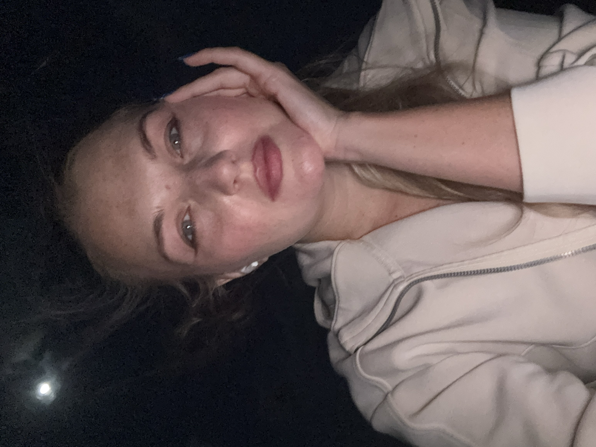 A close-up selfie of a woman is captured at night, with a moon visible in the upper left corner of the dark sky. The woman has blonde hair, fair skin with freckles, and is wearing a light-colored zip-up hoodie. She is resting her chin on her hand, and her eyes are looking directly at the camera. She appears to be wearing makeup, including eyeliner and lip gloss. Her right hand, with painted blue fingernails, is visible near her face. A white wireless earbud is in her left ear. The background is dark, suggesting an outdoor setting at night. There is no information in the image to indicate the location in Denpasar, Indonesia.