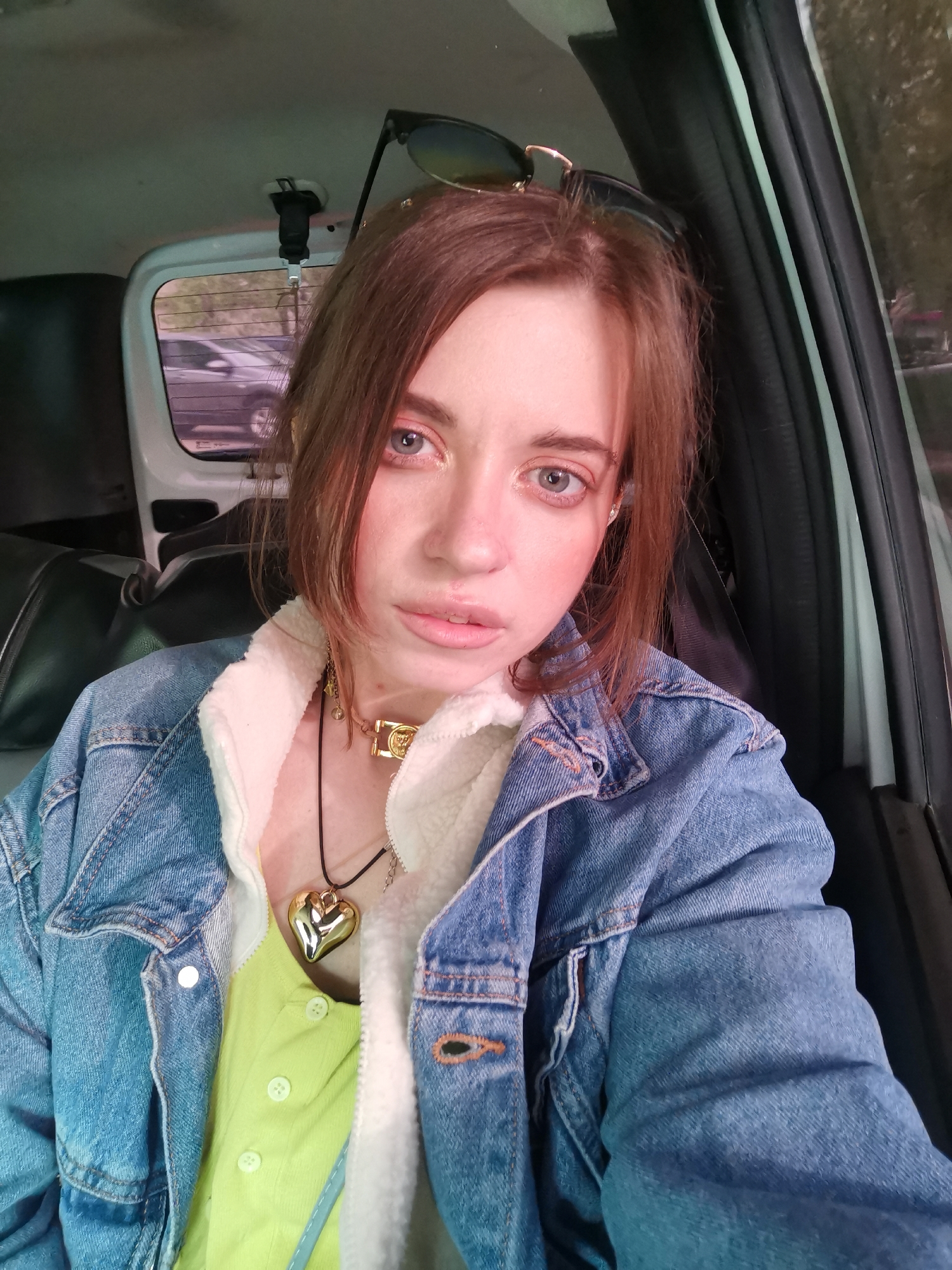 A fair-skinned woman is seated inside a vehicle, facing the camera. She wears a blue denim jacket with white sherpa lining, revealing a bright green shirt underneath. A large gold heart-shaped pendant hangs from a black cord around her neck. She also wears a gold choker-style necklace. Sunglasses are perched atop her reddish-brown hair. The interior of the vehicle is visible, including a black headrest and part of a window with another vehicle visible outside. The location is Unknown, Unknown.