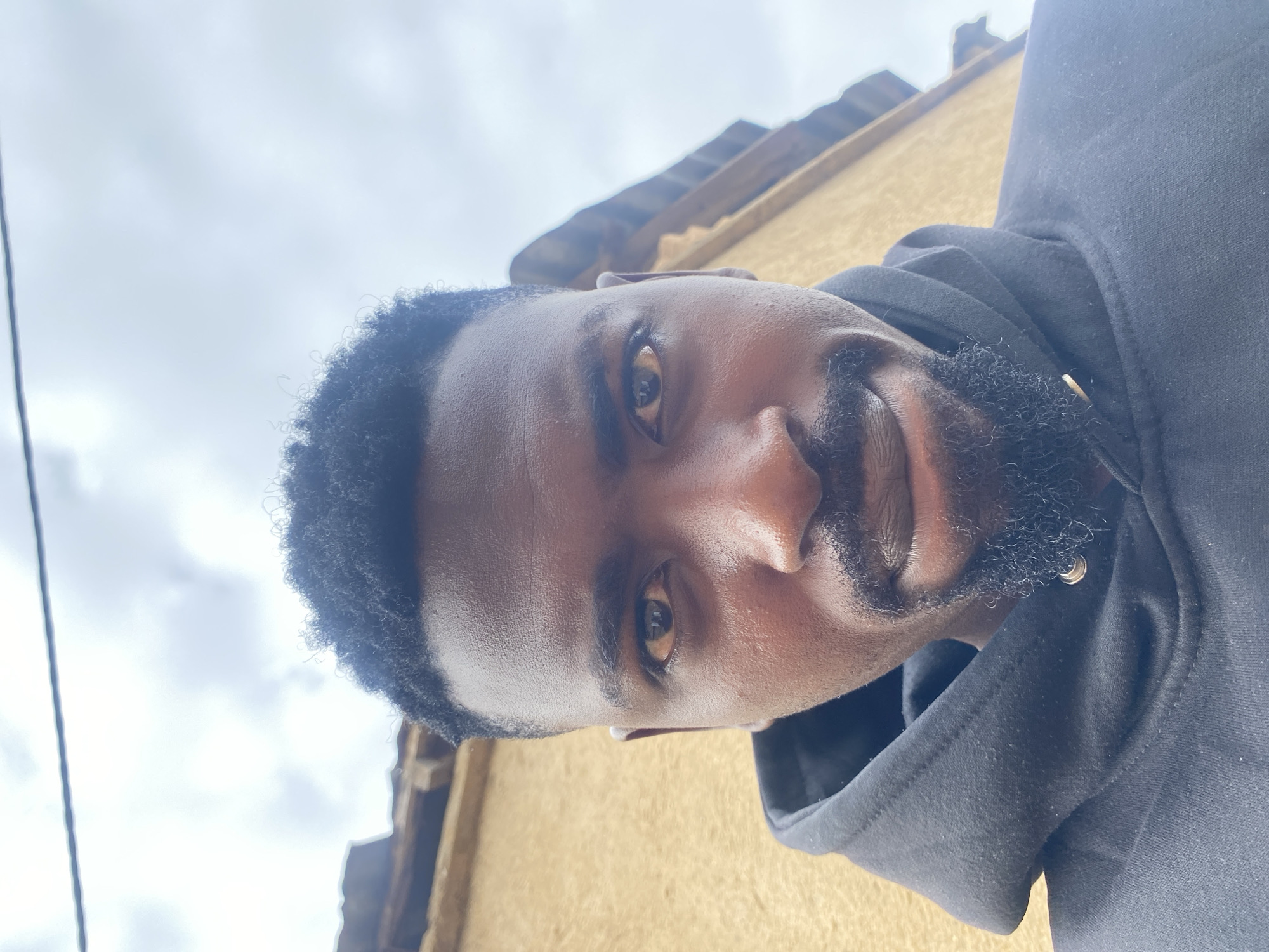 A close-up, upward-angle photograph shows a man with dark skin, short curly black hair, and a trimmed beard and mustache. He is wearing a black hooded sweatshirt. The background includes a light yellow building and a partly cloudy sky. Although no specific location in Barakin Akawo, Nigeria, can be determined based on the image alone, it is inferred the photo was taken there. No further details are discernible about activities or interactions.