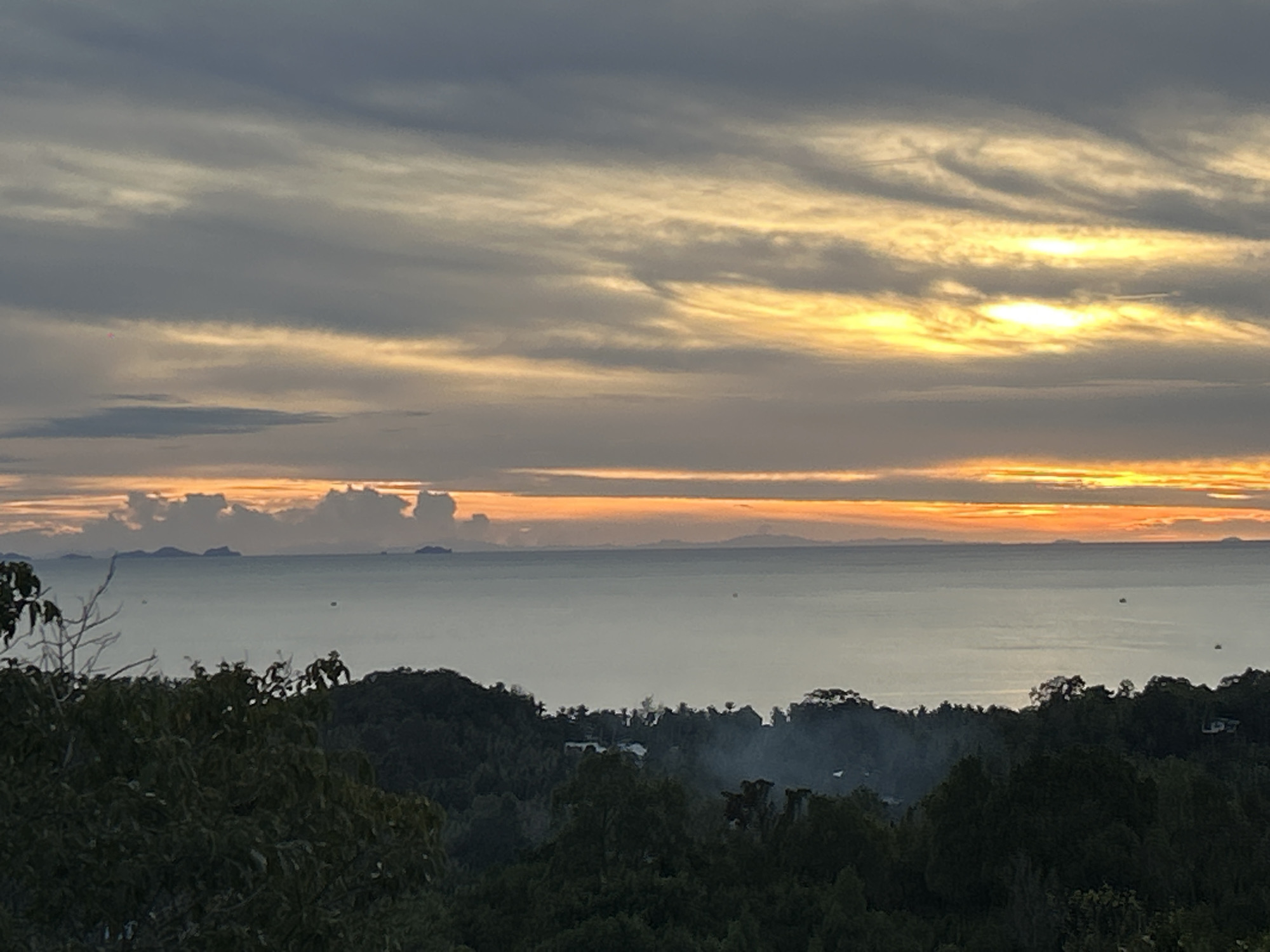 The image presents a scenic view of a sunset over the sea from Baan Hin Kong, Thailand. The sky is filled with layered clouds, transitioning from dark grey to yellow and orange near the horizon, suggesting the sun is setting. Below the horizon line, there are several small islands or landmasses visible across the water. The sea is a hazy grey, with what appears to be small boats or objects scattered across the surface. The foreground is dominated by dark green foliage, indicating a forested area. Some houses can be seen nestled in the trees. There is also smoke rising among the trees.