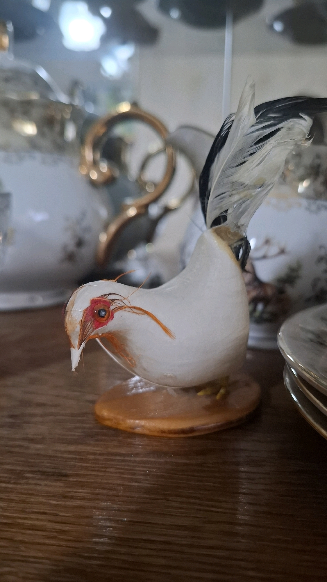 A close-up, eye-level shot captures a decorative ceramic bird with white plumage, a red eye, and black and white tail feathers. The bird sits on a circular orange base, which rests on a wooden surface. The background is blurred, showing hints of a china set with gold accents and glass shelving. The overall scene suggests an indoor setting, possibly a display cabinet or shelf. The location of this image within Shymkent, Kazakhstan, cannot be determined from the visual information provided.