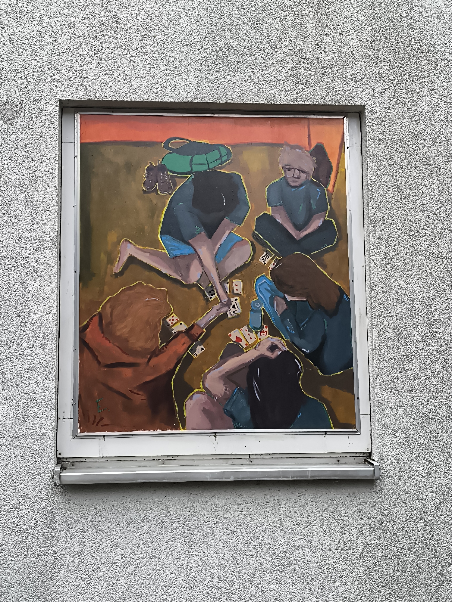 The image shows a painting displayed in a window on a textured grey building wall. The painting depicts five people sitting on the floor, playing cards. The background of the painting is a warm gradient of orange to brown. In the upper left corner of the painting, there are a pair of brown shoes and a green bag. The people are rendered in a somewhat abstract, impressionistic style with bold brushstrokes. One person, wearing an orange top, is in the foreground, their back to the viewer, with their head tilted down towards the cards. Other figures are also engaged in the game, with some looking at their cards and others observing the game. A blue water bottle is visible on the floor. The painting is framed by a white border and installed in what appears to be a window. The building exterior is rough and uneven.
