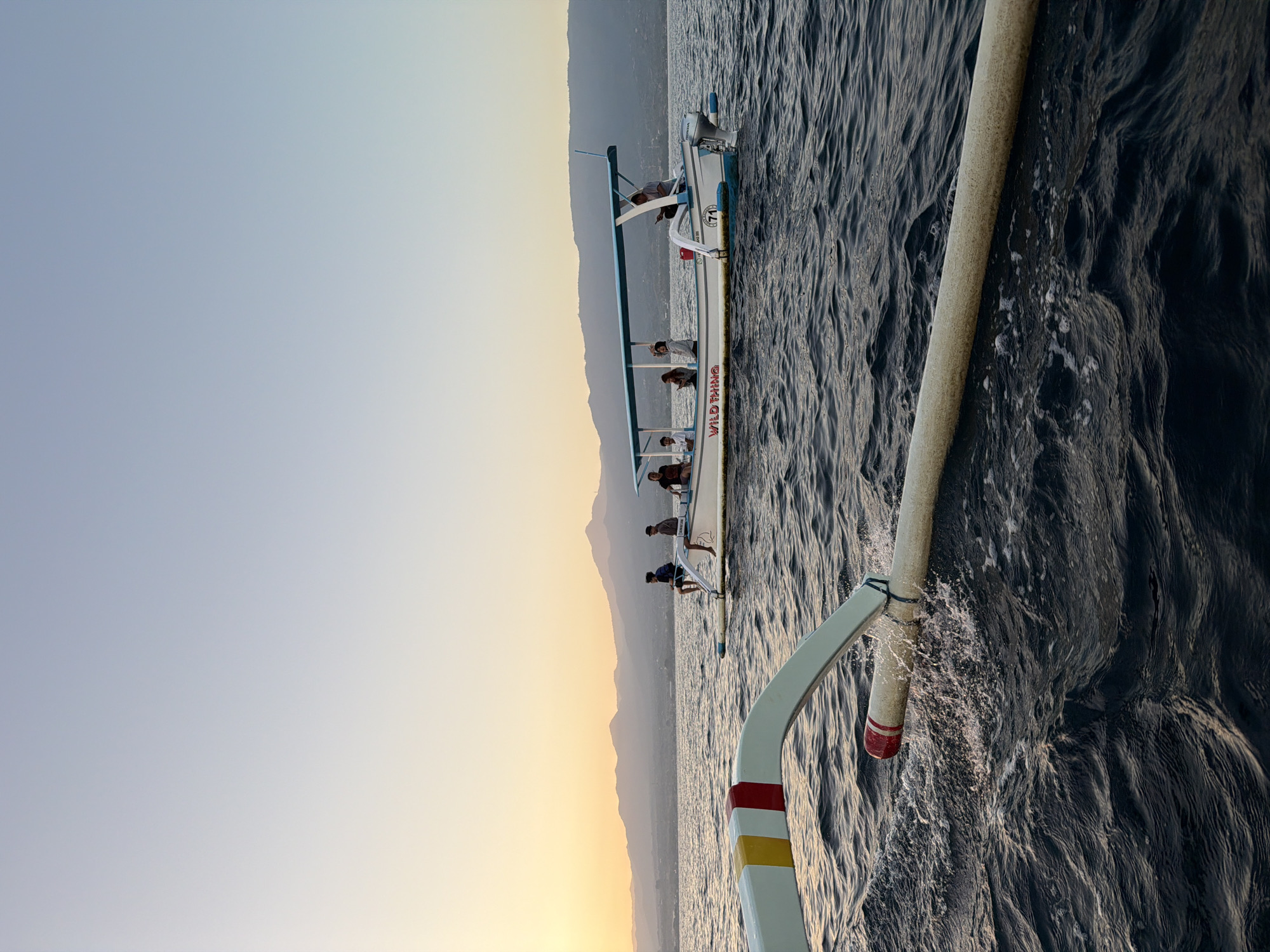 In the city of Buleleng, Indonesia, during sunset, a boat named "WILD THING" is visible on the ocean. Several individuals are seated on the boat, which has a blue canopy. The boat is powered by an outboard motor. In the foreground, another boat is partially visible, with its outriggers extending into the choppy water. Mountains are silhouetted in the background against the fading light of the sky.