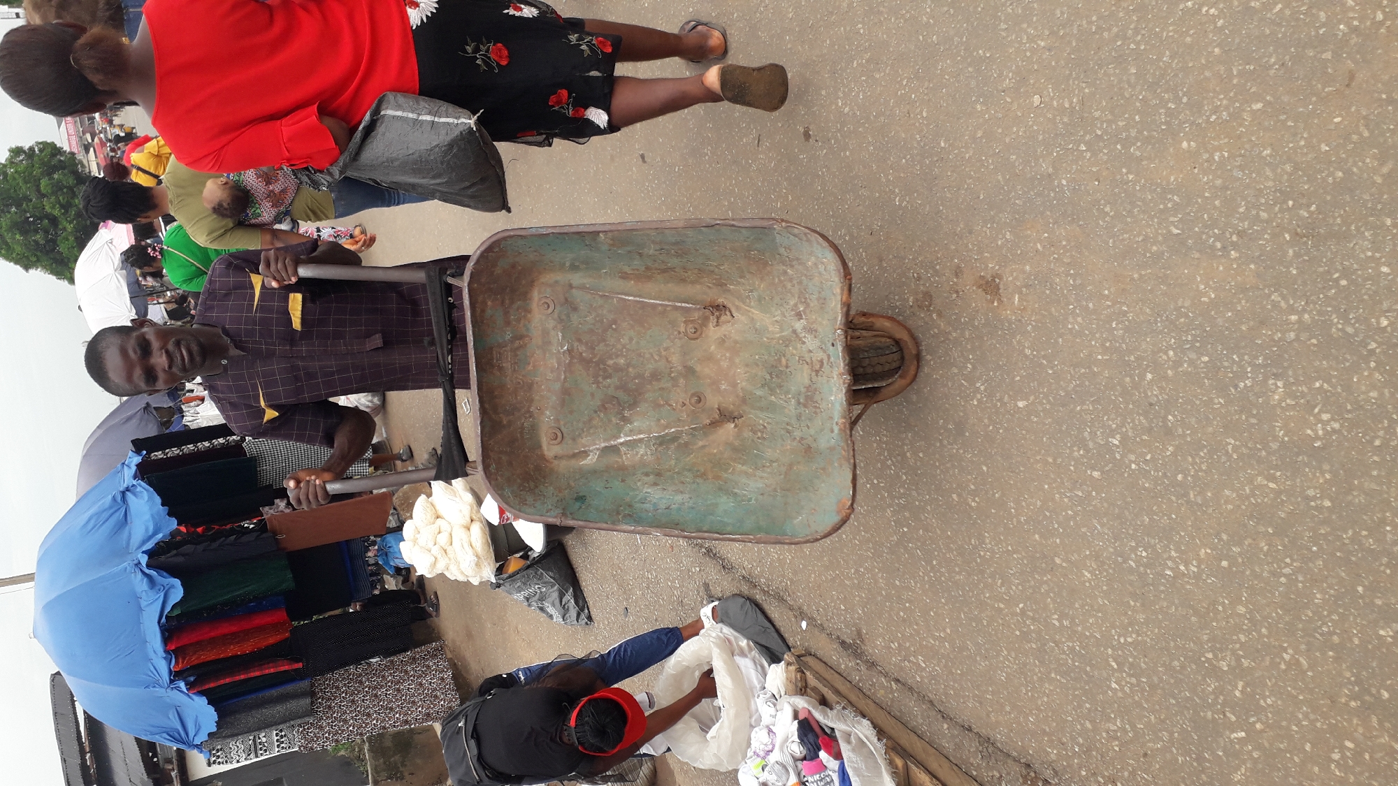 The image depicts an outdoor scene in the city of Onitsha, Nigeria, focusing on a man pulling a rusty, empty wheelbarrow along a busy street. The man is wearing a checkered shirt and holds the wheelbarrow handles. A woman in a black top and red hat is packing white items into a bag next to the wheelbarrow. A blue umbrella is visible, along with colorful fabrics displayed for sale. Other people are walking in the background, including a woman wearing a red top and floral skirt. The ground is paved and shows signs of wear. The overall scene suggests a bustling marketplace environment.