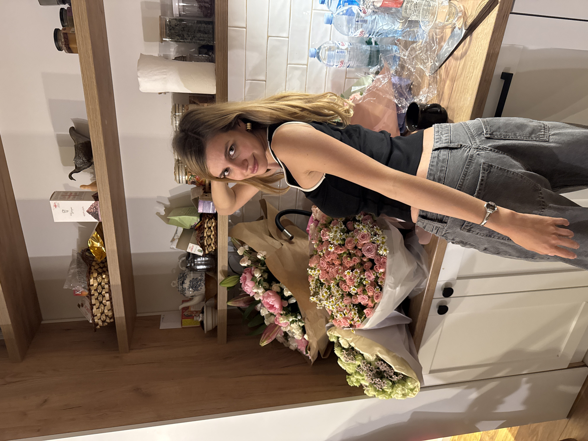 A woman is posing next to a counter with three bouquets of flowers on top of white cabinets. The woman is wearing a black tank top with a white trim and gray jeans. The counter is made of wood. On the counter is water bottles and a black mug. The background is a wall with white tiles and a wooden shelf. The shelf is filled with various items. The location appears to be inside a house.