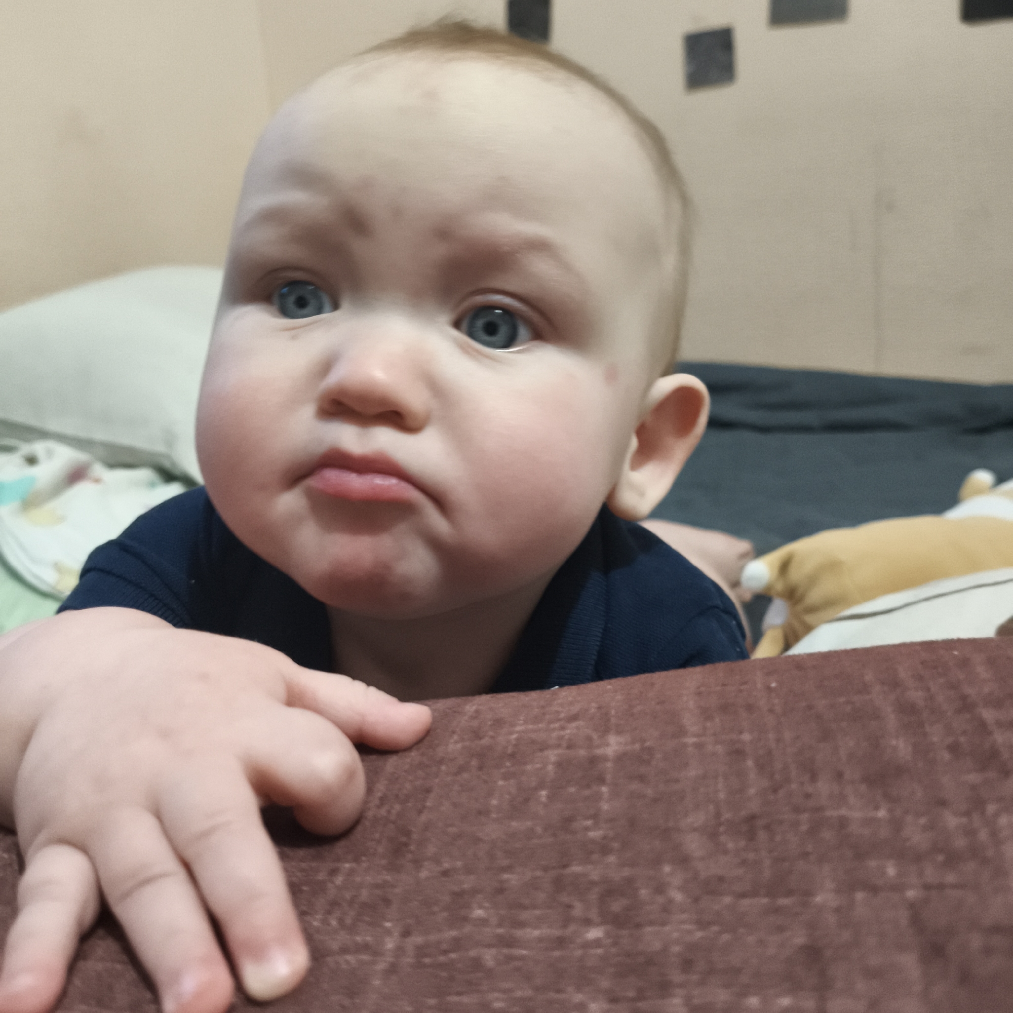 A close-up shot depicts a baby with a pouty expression. The baby has fair skin, blue eyes, and sparse reddish hair. The baby is wearing a dark blue shirt and is resting its hand on a brown, textured surface. In the background, there is a pillow with a patterned cover, a dark blue surface, and a stuffed animal. The background also includes a beige wall with dark, square shapes on it. The image does not provide any information about a city.