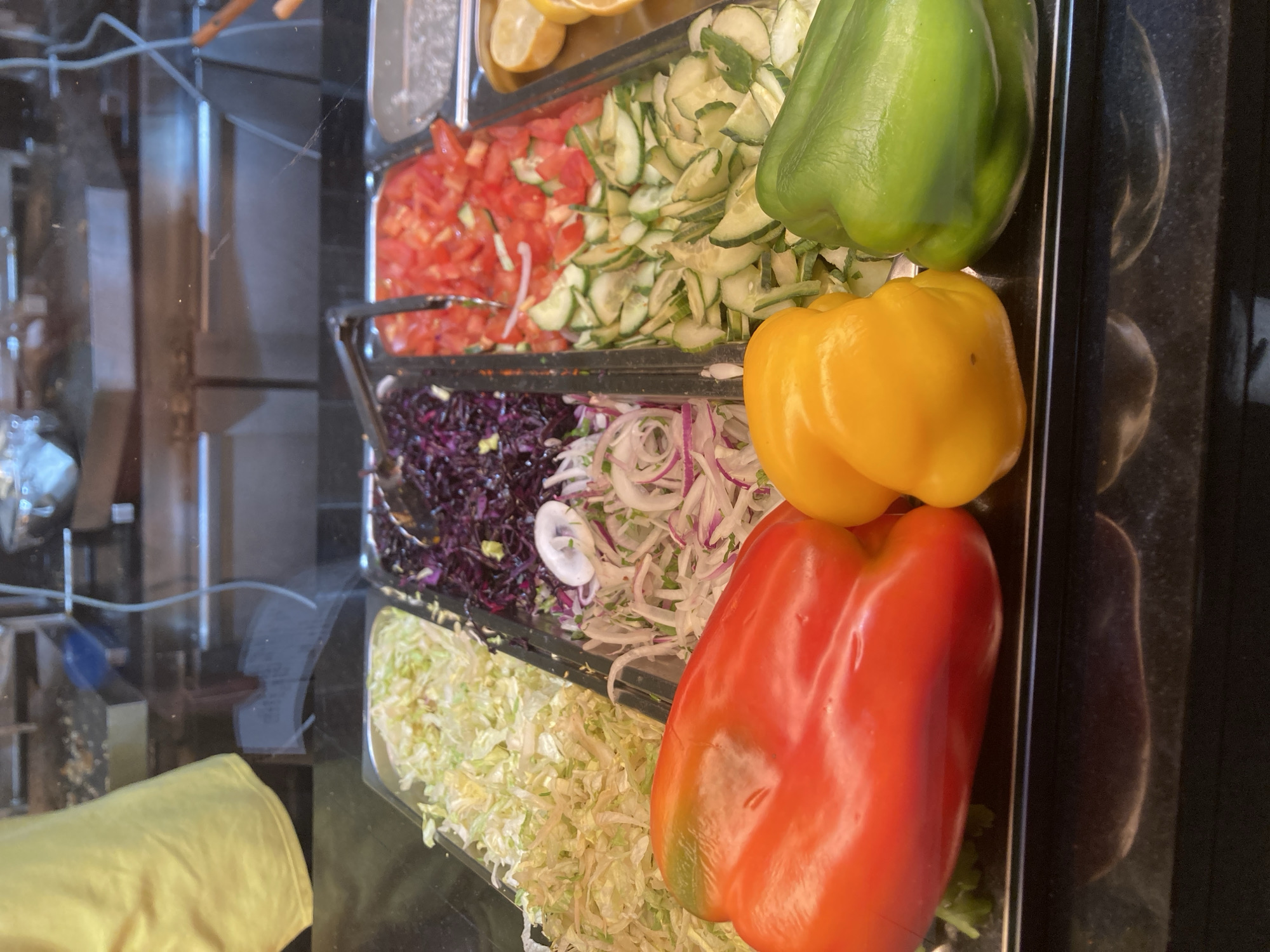 This is a close-up, eye-level, medium shot of a food display in what appears to be a restaurant or food stall. The foreground is dominated by three large, whole bell peppers: a red one on the left, a yellow one in the center, and a green one on the right. Behind the peppers, in a stainless steel tray, are various chopped ingredients: shredded white cabbage on the far left, followed by thinly sliced red cabbage, then a mix of finely chopped red onion and parsley, and sliced cucumbers and tomatoes. Further back, partially visible, are lemon wedges. The background shows kitchen equipment and a yellow apron hanging, suggesting a commercial food preparation environment. The image is brightly lit, likely from overhead sources, and the reflective surface of the display case and the counter creates some glare and distortion. There is no visible signage or context that would specifically place this scene in Barcelona, Spain.