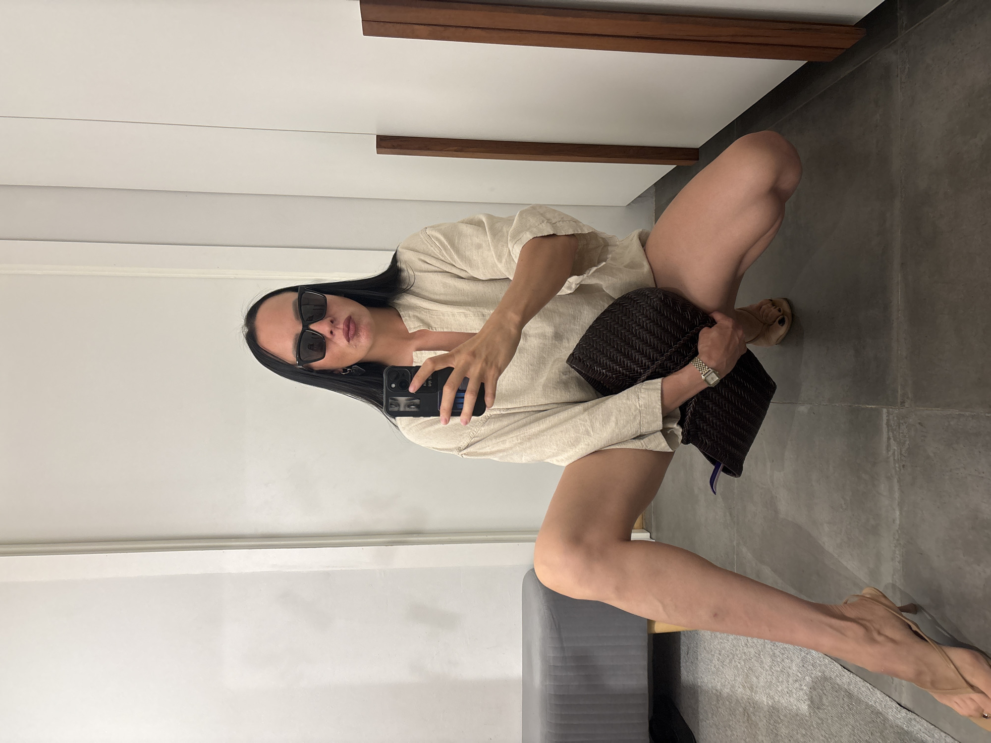 A woman in a neutral-colored linen shirt, sunglasses, and beige heels is squatting and taking a selfie with a black smartphone. She is holding a dark brown woven handbag in one hand. The setting appears to be indoors, likely a room in a home or apartment. The background includes a white wall and a grey tiled floor. Part of a grey upholstered bench or sofa is visible in the lower left corner. The location in the city of Chaikovsky, Russia is not evident from the image.