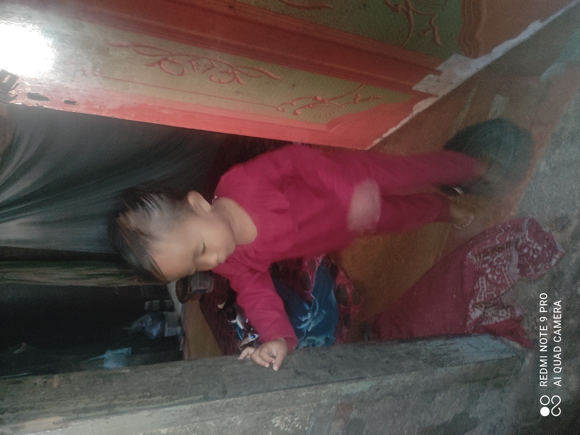 A young child, likely a toddler, is standing in what appears to be a doorway or alcove. The child is wearing a bright red long-sleeved shirt and matching pants. Their hair is dark and slicked back. The child is looking to their left, with their left hand extended and touching a textured, weathered wooden surface that forms the side of the doorway. To the child's right, there is a reddish-orange vertical structure with ornate green and red painted designs, possibly a doorframe or decorative panel. The background is dim and indistinct, with hints of fabric and what might be stored items. The floor appears to be covered with a patterned textile, predominantly in shades of red and white. The overall lighting is dim, with some brighter illumination on the right side of the image. The image contains a watermark at the bottom left corner indicating "REDMI NOTE 9 PRO AI QUAD CAMERA." The location is identified as Sakra, Indonesia.