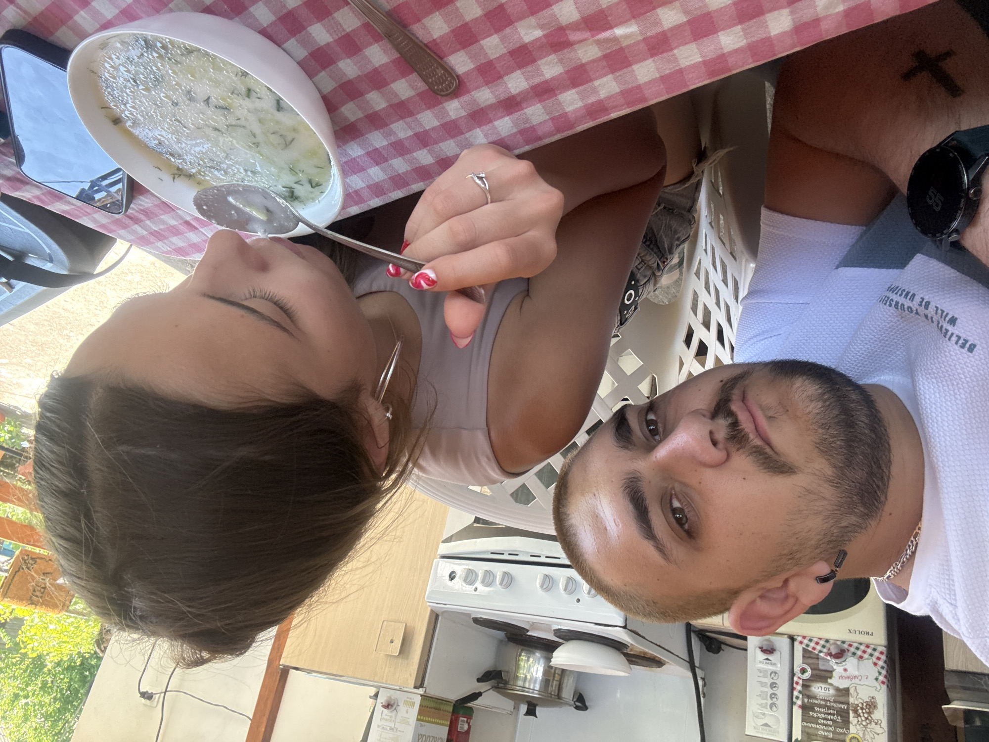 The image depicts two individuals, a male and a female, seated at a checkered table in what appears to be a semi-outdoor kitchen and dining area in Staro Selo, Bulgaria.
The male, positioned on the left, has short blonde hair, a beard, and a mustache. He wears a white textured polo shirt, a gold chain necklace, and a black razor blade-shaped earring in his left ear. A black smartwatch is visible on his left wrist, along with a dark cross tattoo on his left forearm. He is looking directly towards the camera.
The female, positioned on the right, has dark brown hair pulled back. She wears a light-colored sleeveless top and grey denim shorts are partially visible beneath the table. She has silver hoop earrings in her left ear, and a silver ring with a small charm on her left ring finger. Her fingernails are painted with a red and white design. She is actively eating, holding a silver spoon with food to her mouth from a white oval bowl containing a light-colored soup with green herbs, consistent with Tarator. A black smartphone is resting face-up on the tablecloth to the right of her bowl.
The table is covered with a red and white checkered tablecloth. In the background, a kitchen area is visible with a white two-burner electric stove, a pot, and a white bowl on it. A white PROLUX microwave oven is situated to the left of the stove. Above the microwave, two cardboard packages with Bulgarian text are visible, one featuring grapes and mentioning "Thracian wine" ("тракийска вино") and the other mentioning "Bulgarian garlic" ("български чесън"). To the right, a wooden cabinet or door frame is present, along with an electrical outlet and cable. Beyond the dining area, lush green foliage and bright sunlight indicate an outdoor setting during daytime, with a wooden railing partially visible. White plastic chairs with a lattice backrest are also partially visible.