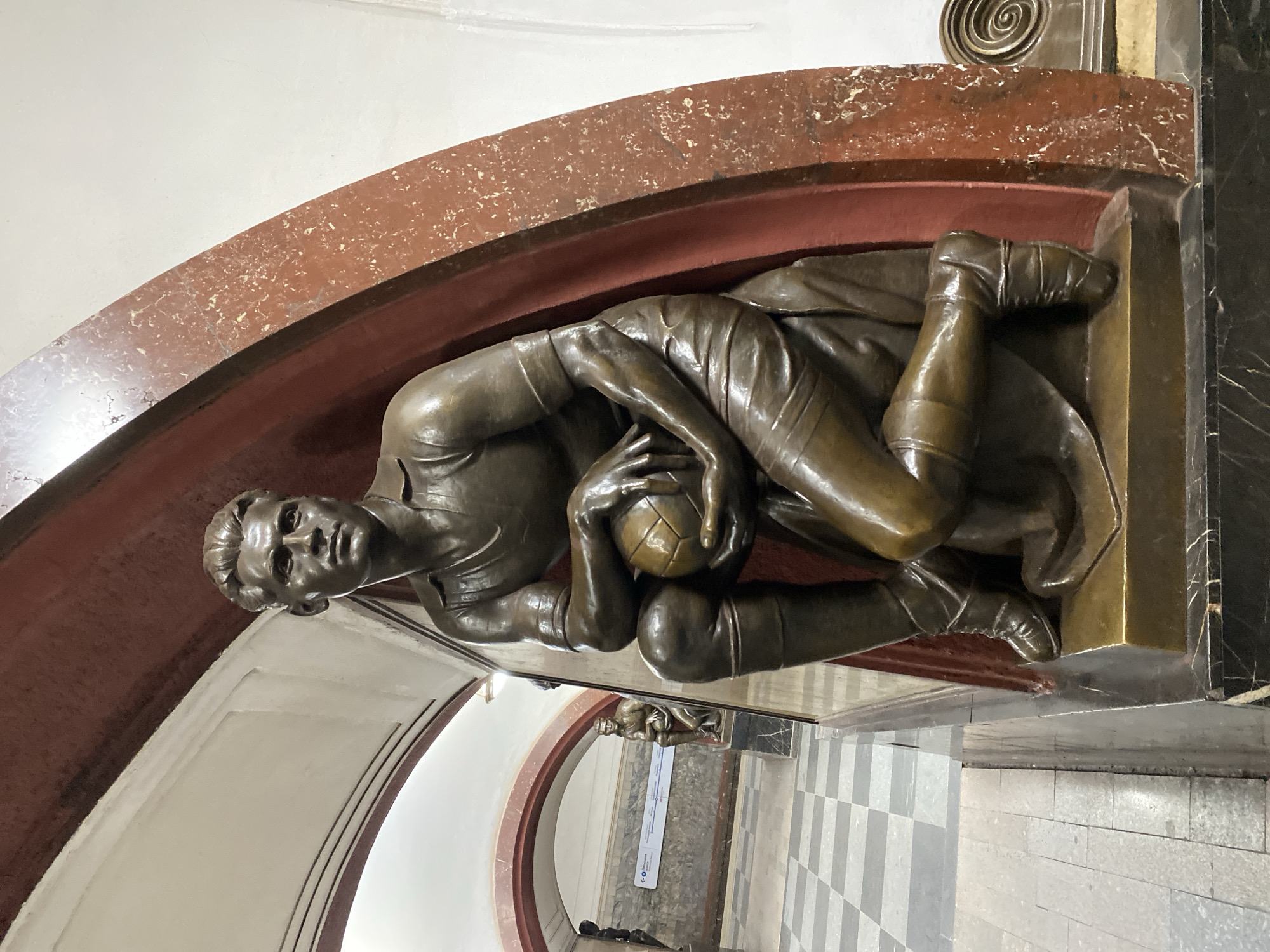 The image depicts a bronze sculpture of a soccer player, lying down and holding a soccer ball, located in the Dynamo metro station in Moscow, Russia. The sculpture is recessed into a wall with a red marble arch above it. The station's interior is visible, including checkered flooring and another archway in the background.