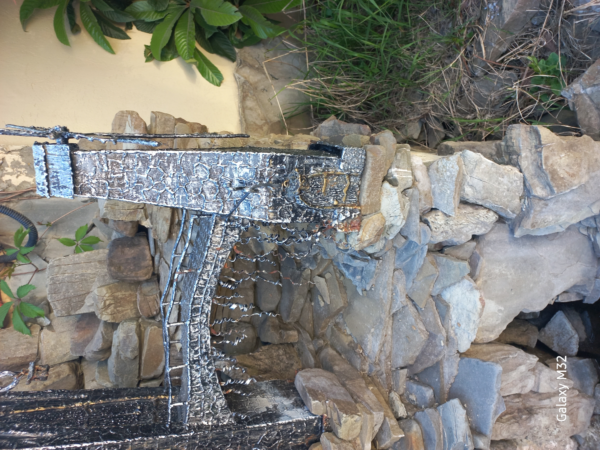 The image depicts a decorative waterfall feature constructed from irregularly shaped rocks and a metal structure. The metal appears to be painted in black and silver, with a textured surface. Water cascades down the metal structure into a rocky basin. The surrounding environment includes vegetation, such as green leaves and grass. The precise location within the city of Sochi, Russia, is not discernible from the image alone.