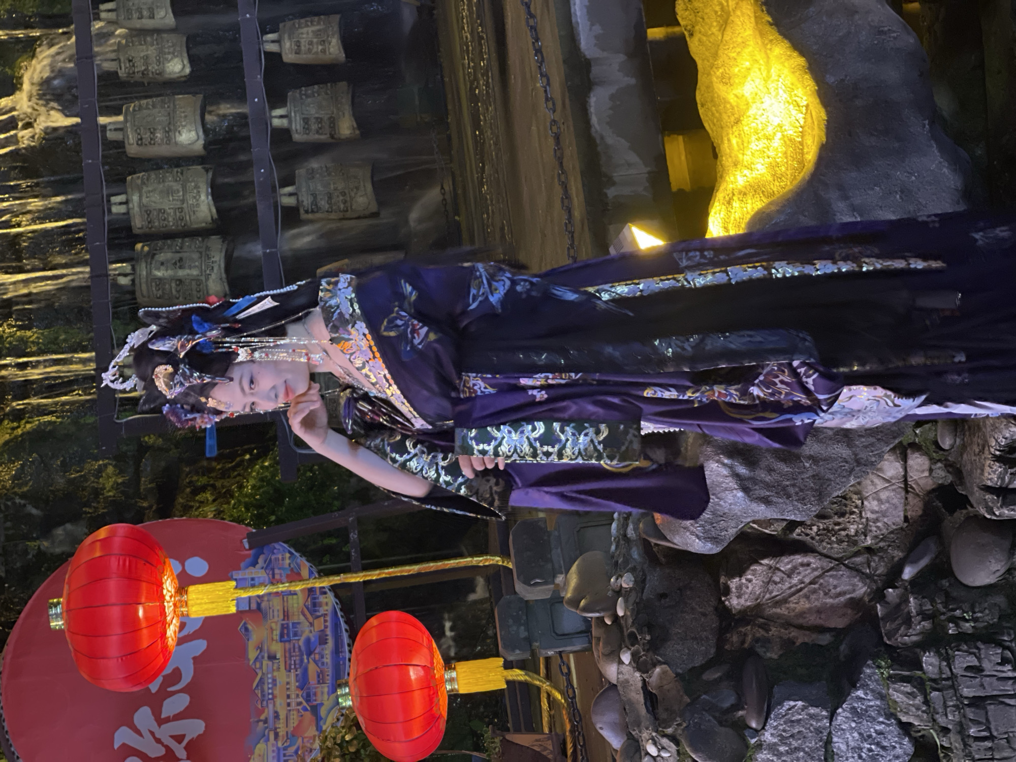 The scene depicts a female individual situated in Yuzhong District, China, under artificial illumination at night. She stands centrally, her body angled slightly left, while her head is turned towards the viewer, displaying a slight smile. Her dark hair is styled elaborately, adorned with a prominent headdress featuring intricate metalwork, jewels, and dangling elements. She is dressed in a dark purple, long-sleeved garment, characterized by extensive gold and silver embroidered patterns. To the subject's left, two illuminated red Chinese lanterns with yellow tassels are positioned, in front of a large red circular backdrop featuring a cityscape illustration and partial white Chinese characters, including '渝'. To the subject's right and extending into the background, a dark, multi-tiered structure supports numerous antique-style bronze bells, behind which water cascades, forming a vertical water feature. A dark walkway with a metal chain barrier crosses the mid-ground. The lower foreground consists of irregular rock formations, with specific areas brightly highlighted by a strong yellow light source.