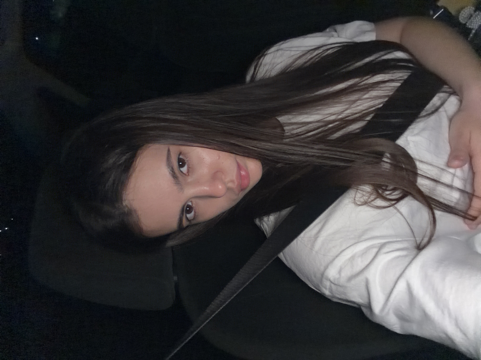 A young woman with long, dark hair is seated in the driver's seat of a car at night. She is wearing a white t-shirt and her seatbelt is fastened across her chest. The background is dark, suggesting the car is moving or parked in a dimly lit area. Her facial expression is neutral, and her gaze is directed towards the camera. There is no visible indication of the location within the city of Chaikovsky, Russia, or any specific environmental details to suggest it. The image captures a personal moment, likely a selfie, taken inside the vehicle.
