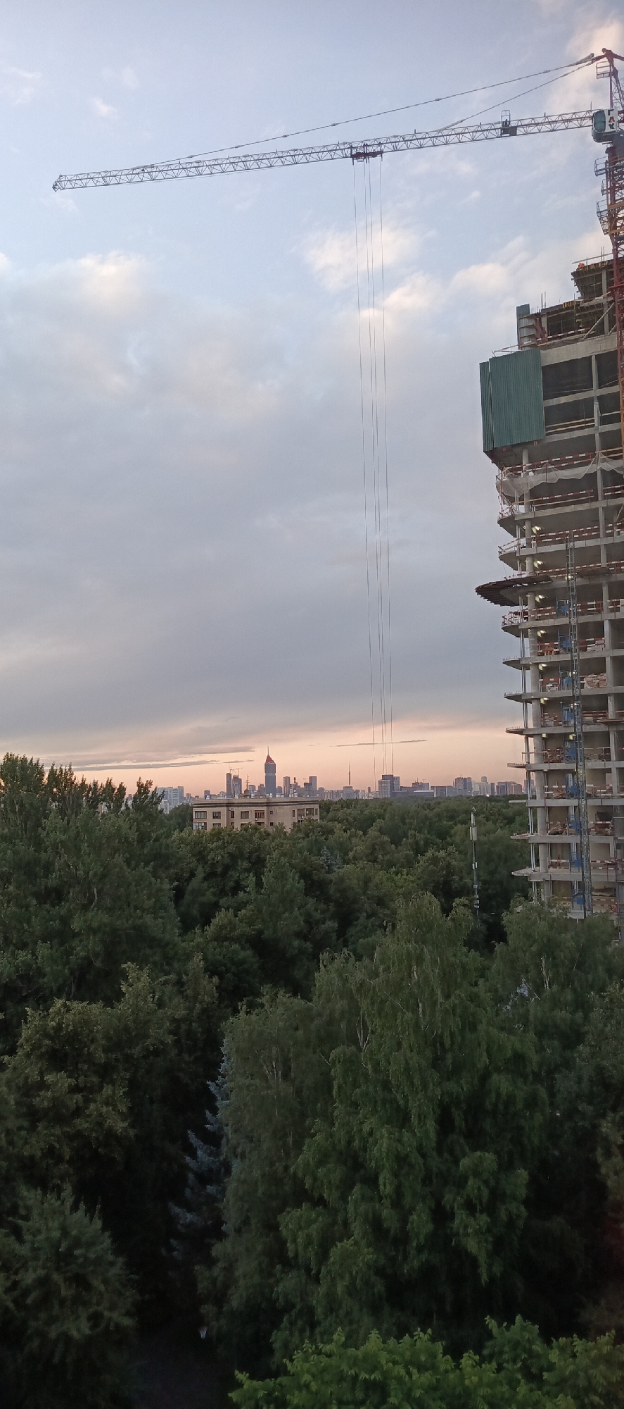 This image captures a construction site in Moscow, Russia, at dusk. The primary subjects are a large, partially constructed building and a towering crane that stretches across the sky. The building, made of concrete and scaffolding, is in the foreground on the right, with multiple floors visible and construction materials piled on some levels. A green protective sheeting is attached to one side of the building. The crane, a prominent object, has a long horizontal jib extending from its main tower, with several cables hanging down.
In the background, a dense forest occupies the mid-ground, separating the construction site from a distant city skyline. The skyline features several skyscrapers, one with a distinctive red spire, indicative of a major urban center. A low-rise building with a light-colored facade is visible within the treeline, just before the skyscrapers.
The sky is filled with soft, diffused clouds, with hues of pale blue, gray, and a hint of pink near the horizon, suggesting the sun has recently set. The overall mood is serene, with the quiet of dusk contrasting with the industrial activity implied by the construction. There are no visible people in the image. The scene is set in an urban environment on the outskirts of Moscow, where development meets natural landscape. No text is visible in the image.