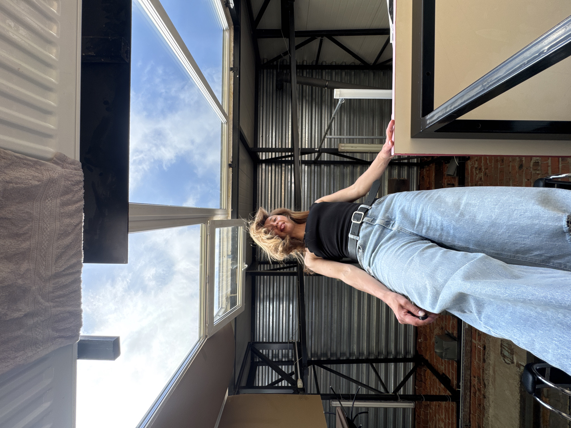 A female stands inside a structure with corrugated metal walls and exposed metal beams, possibly a warehouse or industrial building. She wears a black tank top and light blue jeans with a black belt. She appears to be holding onto a large framed object, likely a piece of artwork. Windows open to the sky, which is lightly cloudy. The location is Chaikovsky, Russia.