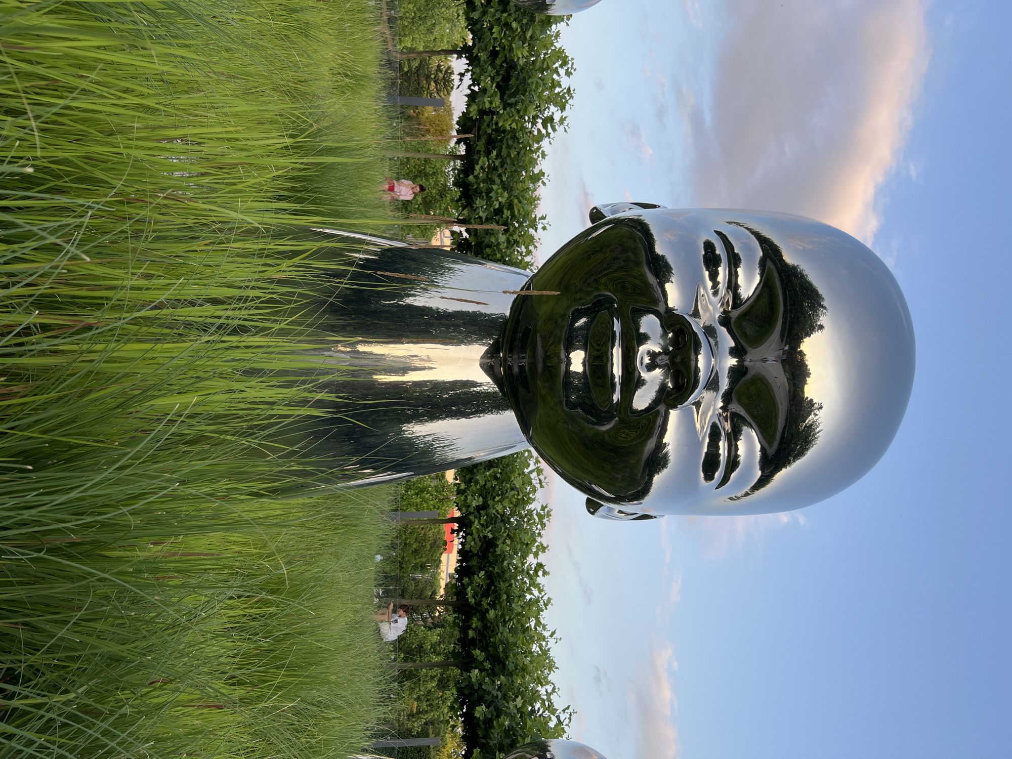 The photograph features a large, highly reflective metallic sculpture of a baby's head in a park setting. The sculpture is positioned in the foreground, surrounded by tall green grass. In the background, there is a line of trees, and beyond that, a clear blue sky with some soft clouds.
The sculpture's polished surface reflects the sky, trees, and surrounding environment, creating distorted images within its sheen. The sculpture appears to be part of a larger installation, as another similar sculpture is visible to the left.
In the mid-ground, to the left, a young person wearing a pink shirt and red shorts is standing and looking towards the sculpture. To the right, a person in a white shirt and shorts is sitting on the grass, looking down at something in their hands.
The location of this sculpture is in the "Park of the First President" in Krasnodar, Russia. The park is known for its contemporary art installations, including the "Babies" sculptures by artist Hossein Namin. The time of day appears to be late afternoon or early evening, given the soft lighting and the colors in the sky. The environment is natural and well-maintained, suggesting a public park or garden.