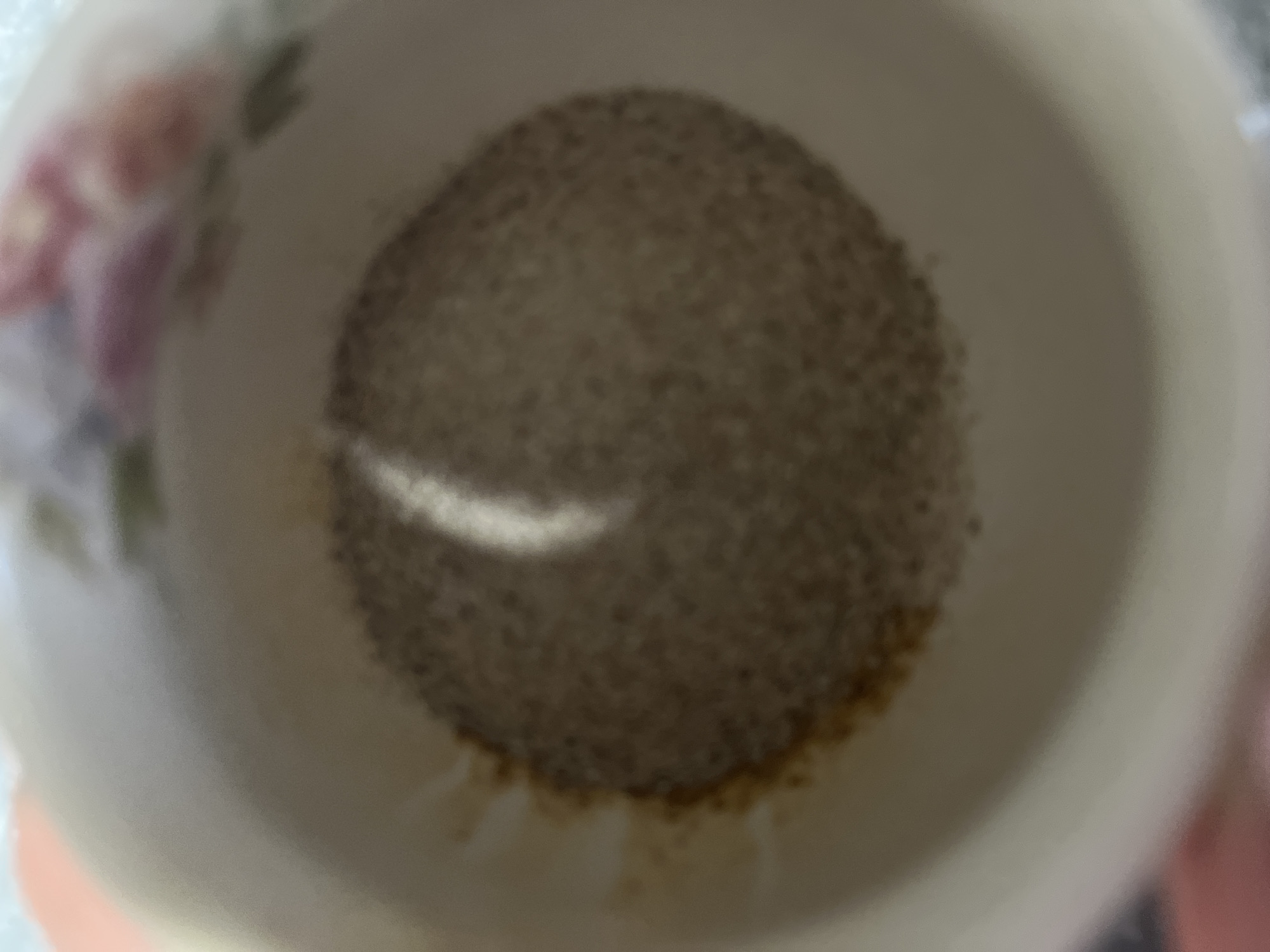 The image is a close-up, high-angle shot looking down into a white ceramic mug. The mug has a floral pattern on its upper rim, with visible hints of pink and green. The bottom of the mug contains a layer of fine, granular material, appearing to be coffee grounds or a similar substance. A bright, vertical reflection of light is visible on the surface of the grounds. The surrounding area of the mug is a pale, off-white color. The image does not provide any specific details about the location in Vladikavkaz, Russia, other than it is a scene depicting the inside of a mug. There are no people, actions, or interactions depicted.