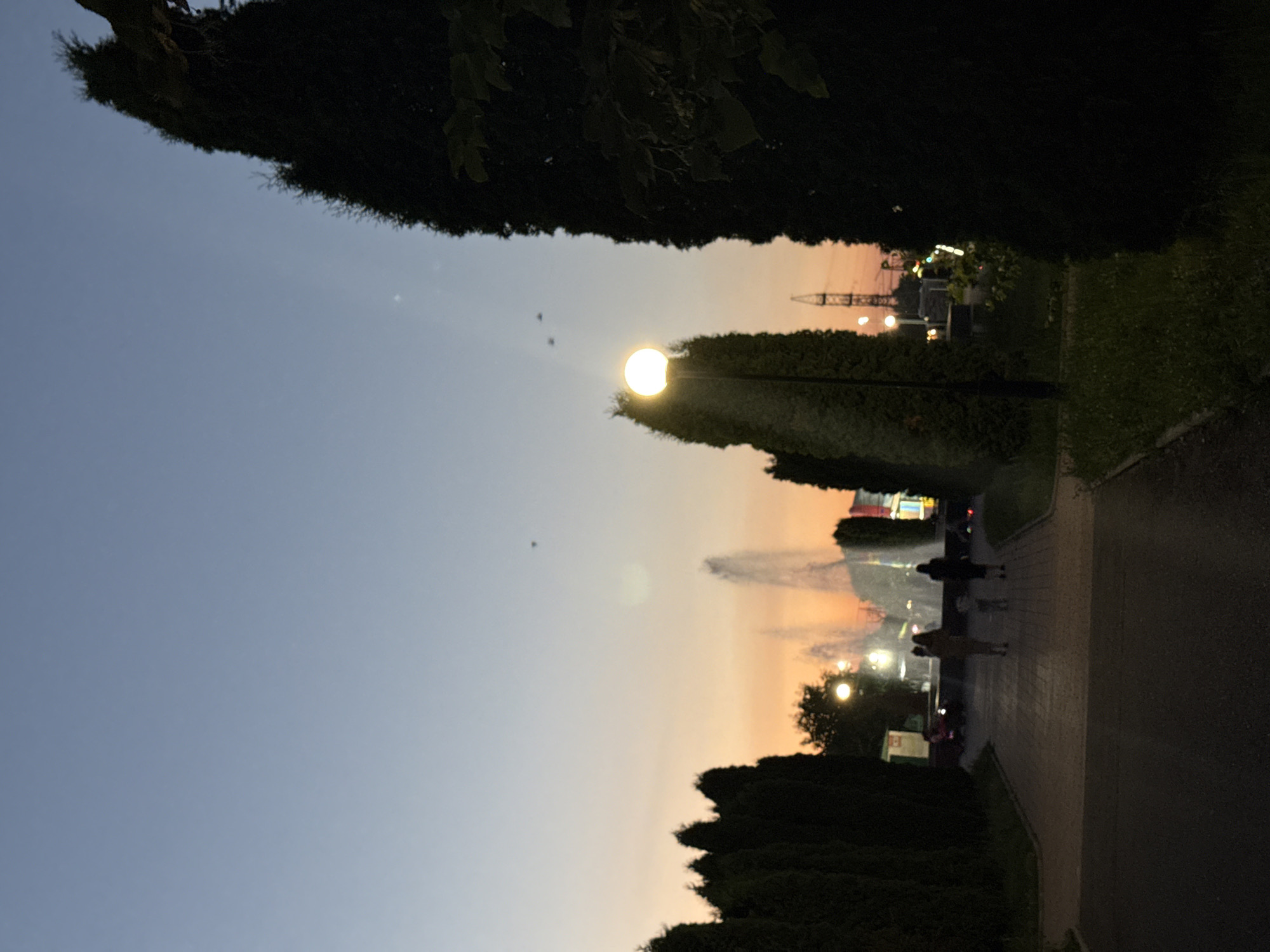 The image depicts a park scene at dusk in Endirei, Russia. A pathway runs through the park, bordered by manicured hedges and grass. Several people are visible walking along the path, their silhouettes dark against the softening sky. A bright, illuminated lamp post stands out in the foreground, casting a warm glow. In the background, a Ferris wheel and other amusement park rides are lit up, suggesting the park is a place of entertainment. The sky is a gradient of soft blue and orange, indicating the transition from day to night, and the overall atmosphere appears peaceful and serene. There is no visible text in the image.