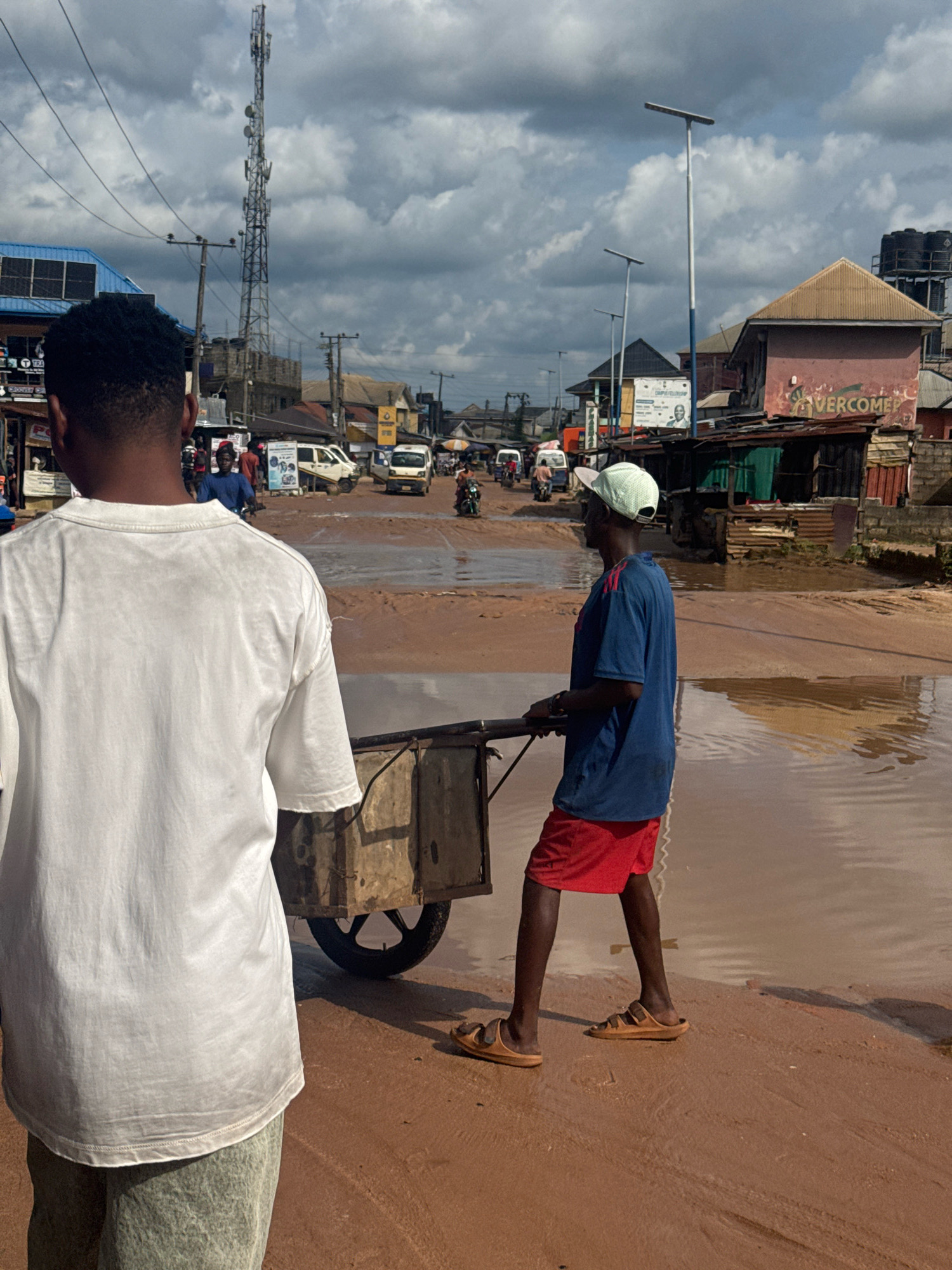 The image captures a scene in Ihiagwa, Nigeria, where a flooded street presents a muddy, watery environment. In the foreground, a person wearing a white t-shirt and light green pants is seen from the back, facing away from the camera. To their right and slightly behind, another person, facing left in profile, is pushing a wooden cart. This individual is wearing a light blue t-shirt, red shorts, a light green cap, and brown sandals. The cart is supported by a single wheel.
The street itself is significantly waterlogged, with the brown, muddy water reflecting the overcast sky and surrounding buildings. In the background, several buildings line the street, some appearing to be shops or residences. A prominent telecommunications tower stands tall against the cloudy sky. Vehicles, including a white van and motorcycles, are visible on the flooded street, suggesting ongoing activity despite the conditions. The overall impression is of a typical urban environment in Nigeria, experiencing weather-related disruptions.