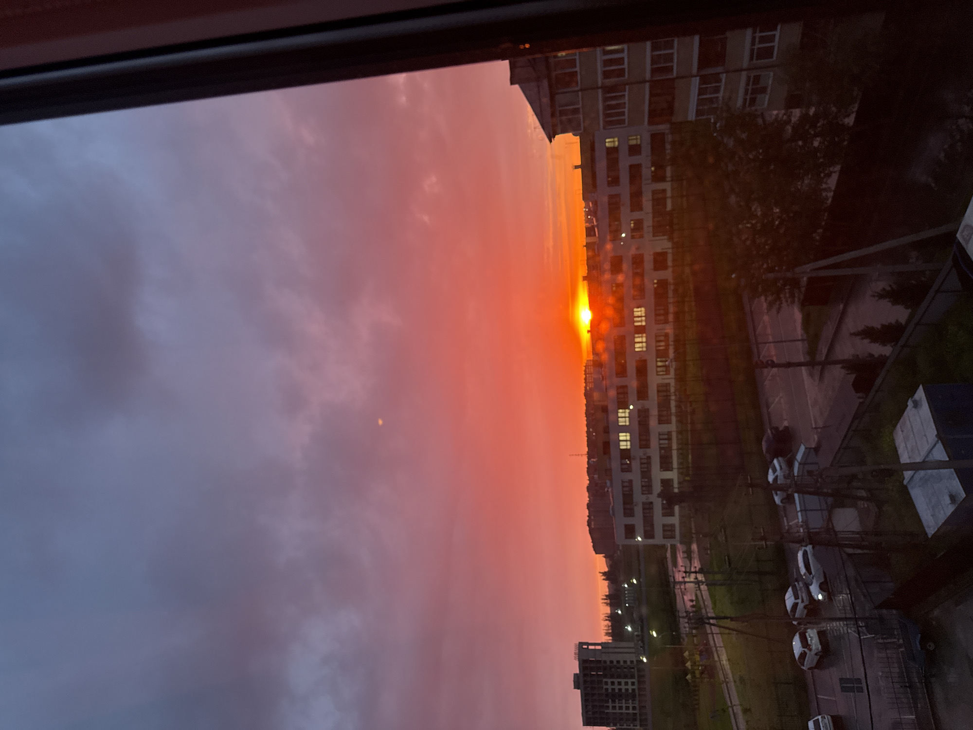 Here's a detailed description of the image:
**Setting:** The image is taken from an elevated position, possibly a high-rise apartment window, overlooking a cityscape during a vibrant sunset. The sky is dominated by hues of purple and red, with the sun appearing as a bright, orange orb on the horizon.
**Objects:**
* **Buildings:** The cityscape includes a mix of high-rise apartment buildings and lower structures. Many have illuminated windows, suggesting it is late in the evening.
* **Streetscape:** Below the buildings, there's a street with cars parked along the roadside. Streetlights are visible, further indicating the time of day. Green patches suggest the presence of parks or landscaped areas.
* **Sky:** The sky is the dominant element, showcasing a spectacular sunset. The clouds are layered, adding texture to the scene.
**Environment:** The scene suggests a calm, urban environment during the twilight hours. The light and colors create a tranquil atmosphere.
**Location:** While the city is unknown, the presence of modern high-rise buildings and streetlights suggests it is a well-developed urban area.
