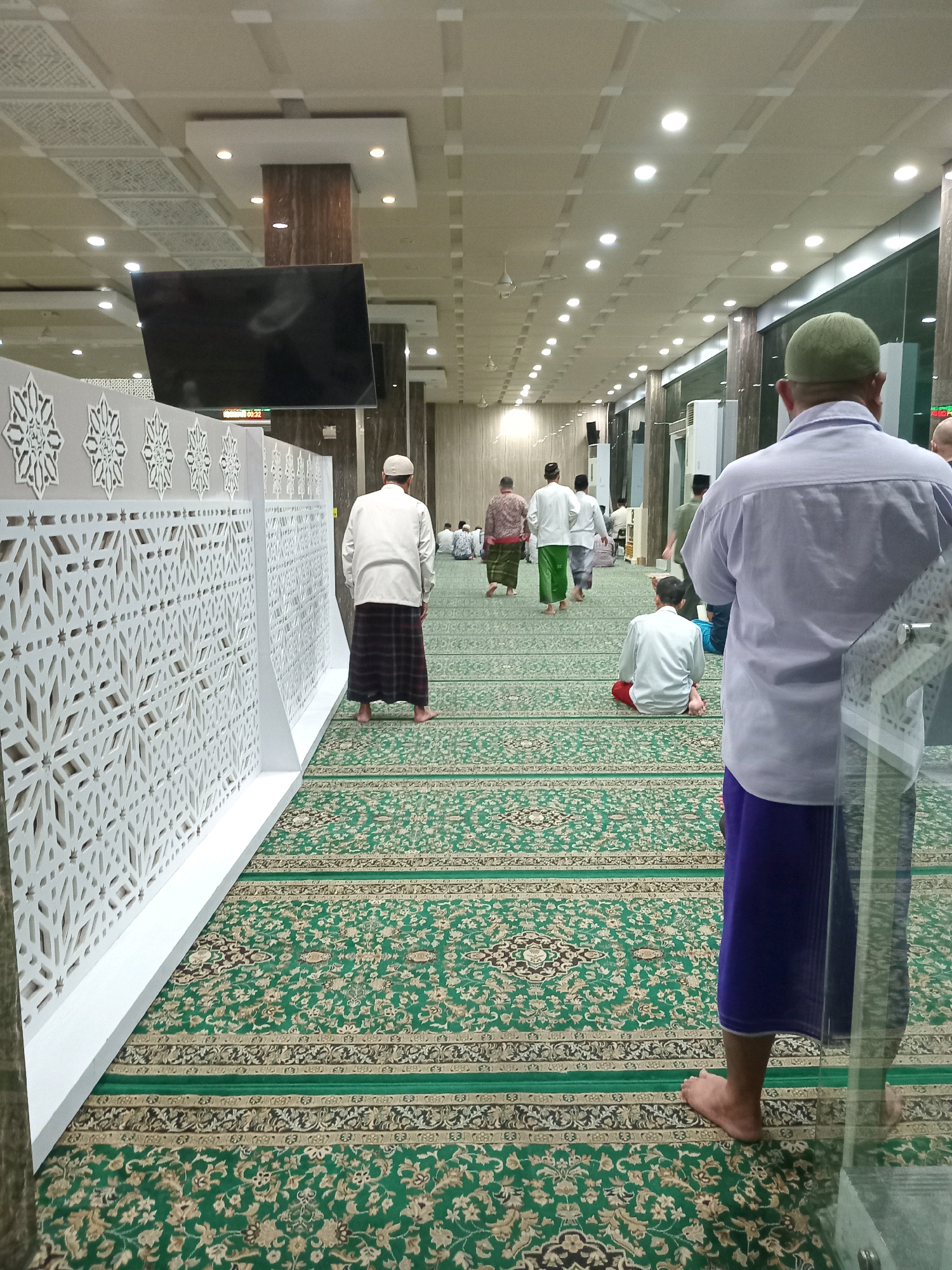 The image depicts the interior of a mosque, specifically the prayer hall, in Randuagung, Indonesia. The scene is dominated by a vibrant green carpet with intricate floral patterns, laid out to define prayer spaces. Several men, dressed in traditional Islamic attire, are visible. Some are standing, facing towards the front of the hall, likely preparing for or observing prayer. Others are seated on the carpet, engaged in prayer or contemplation.
In the foreground, a man wearing a light-colored shirt and a purple sarong is seen from behind, his bare feet on the carpet. To the left, a decorative white partition with a geometric pattern runs along the wall. A large black television screen is mounted on the wall above this partition. Further into the hall, more men are scattered throughout, some walking and others seated. The ceiling is adorned with square panels and illuminated by numerous round lights, creating a well-lit environment. The overall atmosphere suggests a communal religious gathering.