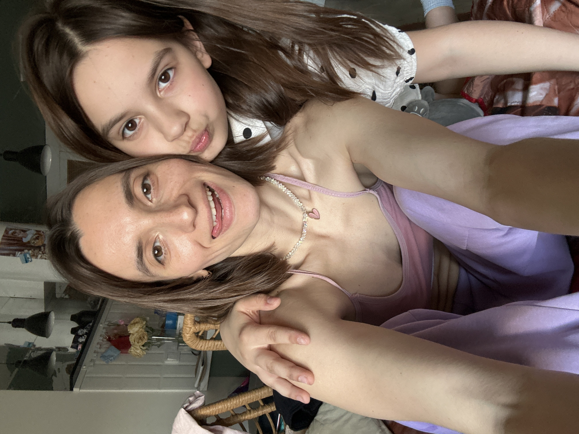 A selfie photograph features a woman and a girl in what appears to be an indoor setting. The woman, with brown hair, is wearing a pink tank top and a necklace with a heart pendant. She is smiling and her tongue is slightly visible. The girl, with brown hair and a polka dot top, is positioned close to the woman and is making a kissing face. A lavender blanket is draped over the woman. A partially visible rattan chair is present. In the background, there is a shelf with various items including flowers, bottles, and a lamp. The location cannot be determined as being Moscow, Russia based on the current image.