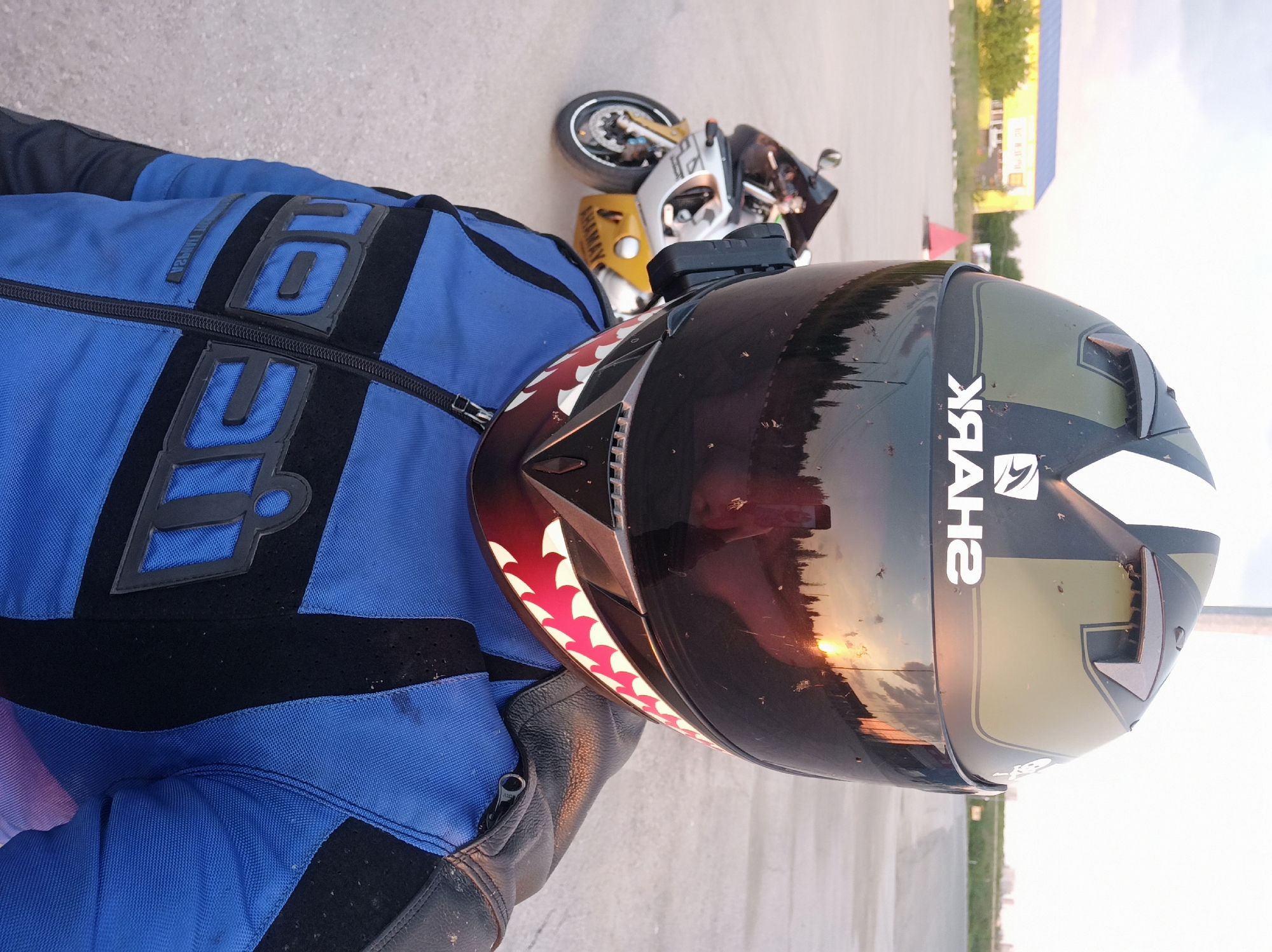 A person wearing a blue and black Icon motorcycle jacket and a Shark helmet with a shark teeth design is looking at the camera. The helmet's visor reflects a sunset or sunrise. A motorcycle is parked in the background. The photo appears to be taken on pavement. The location is Perm, Russia, based on building signage visible in the background, but this may be a guess.