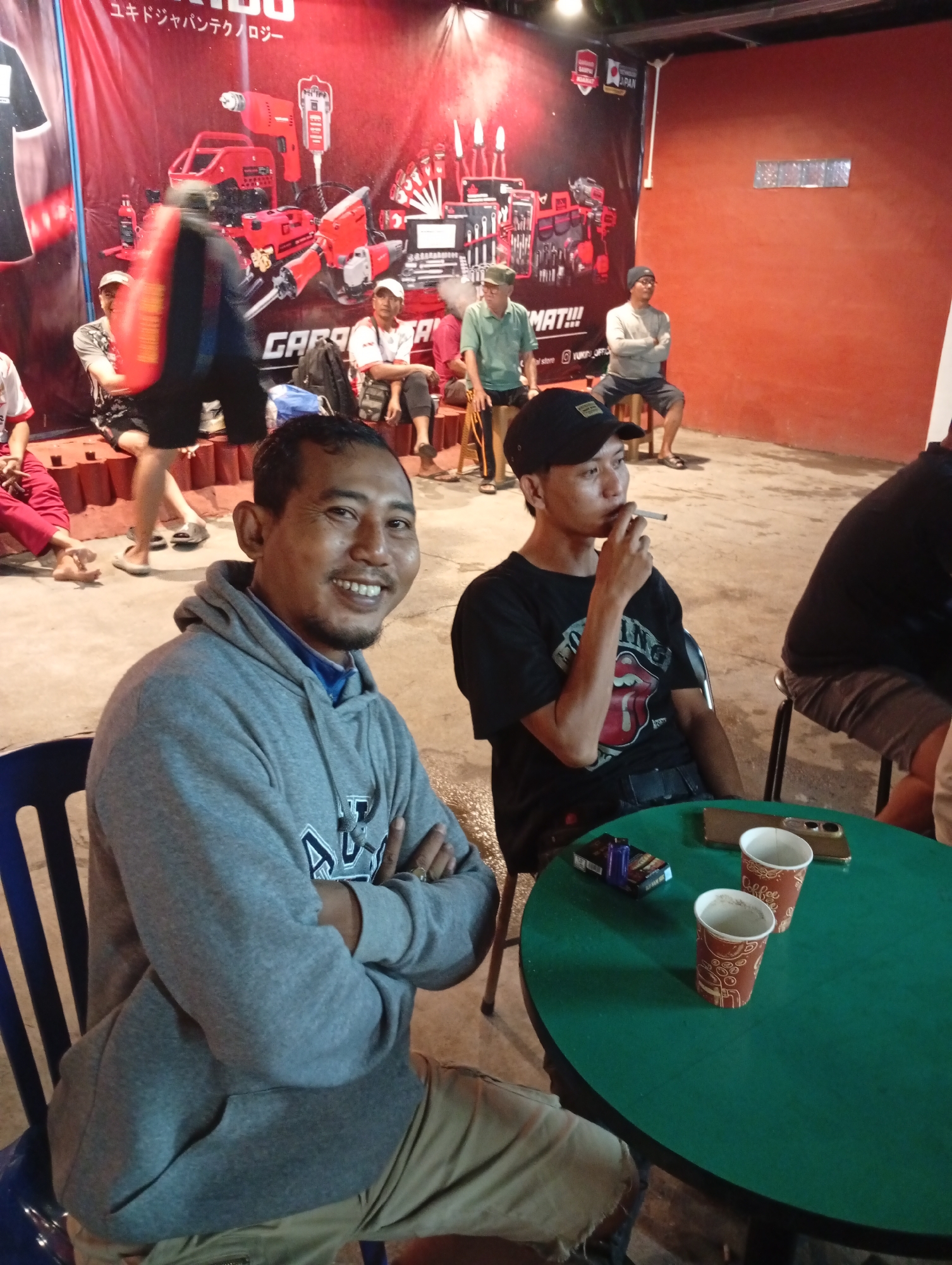 In a dimly lit outdoor setting in Caringin, Indonesia, several individuals are gathered. In the foreground, a man wearing a grey hoodie smiles at the camera, his arms crossed. To his right, another young man in a black t-shirt and cap smokes a cigarette. A green table sits between them, holding two paper cups, a pack of cigarettes, and two cell phones. In the background, a large red banner featuring images of tools and Japanese text hangs on the wall. Several other people are seated on low steps or chairs behind the banner, some blurred by motion. The environment appears to be a casual gathering place, possibly a roadside stall or community area, with concrete flooring and an orange wall visible.