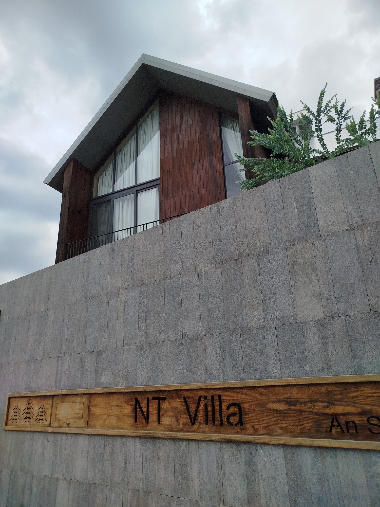 The image presents a low-angle view of "NT Villa An S," located in Da Lat, Vietnam. A two-story wooden house is visible behind a textured gray stone wall. The house's architecture features large windows with white curtains and a simple roofline. To the right of the house, green foliage extends above the wall. A wooden sign with carved lettering displaying the villa's name is affixed to the wall. The sky is overcast. No people are visible in the image.