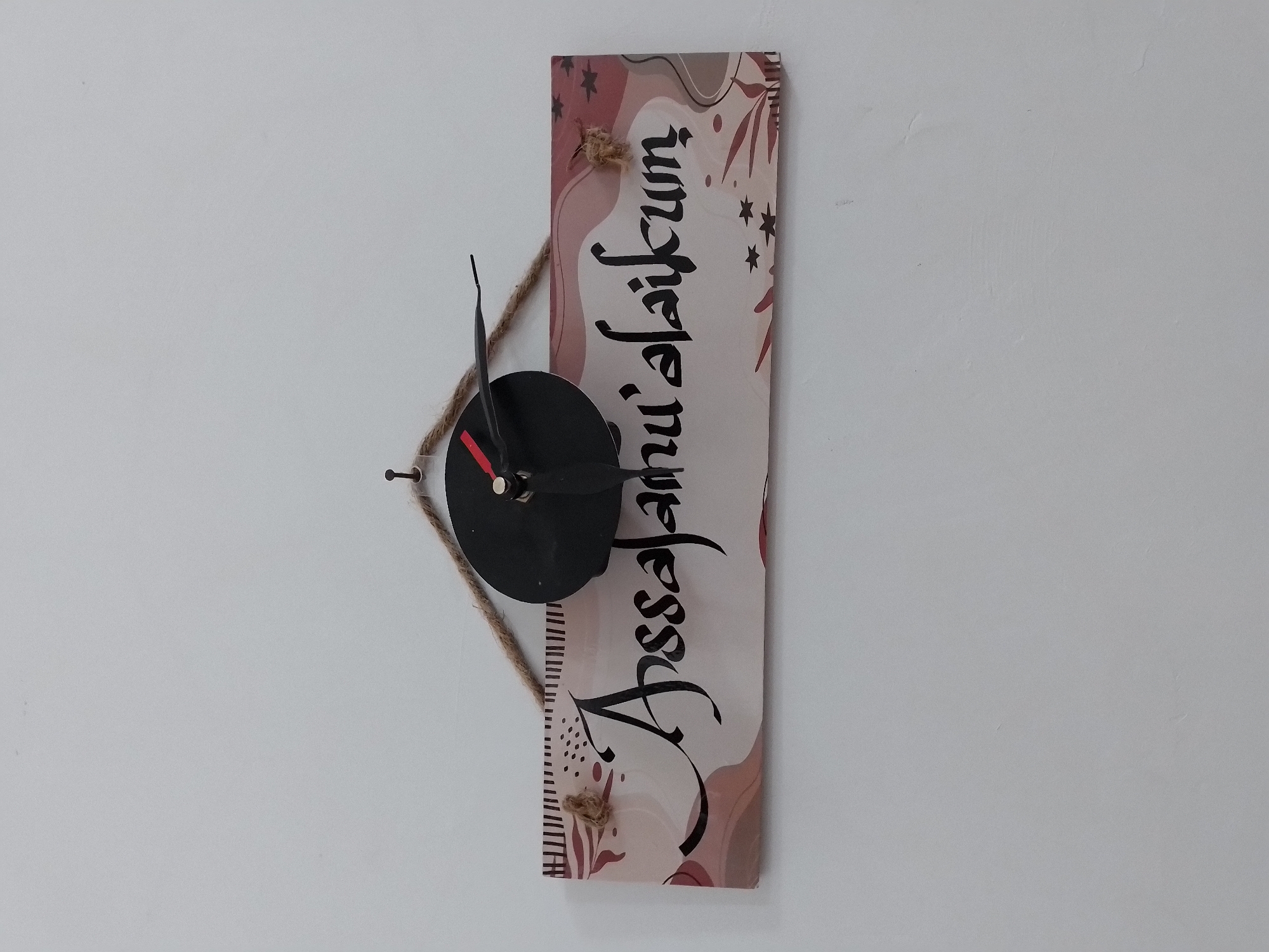 The image shows a wall clock mounted on a white wall. The clock is a decorative piece with a black circular face and black hour and minute hands. It is attached to a rectangular wooden plaque that is decorated with an abstract pattern of pink and brown shapes and small star motifs. Arabic calligraphy, reading "Assalamu'alaikum", is written in black on the plaque. The plaque is hung on the wall using a piece of braided rope, which is tied to two loops on the top edge of the plaque. The clock mechanism and hands protrude from the center of the plaque. The wall appears to be painted white and has a textured surface. The image was taken in the city of Ngancar, Indonesia.