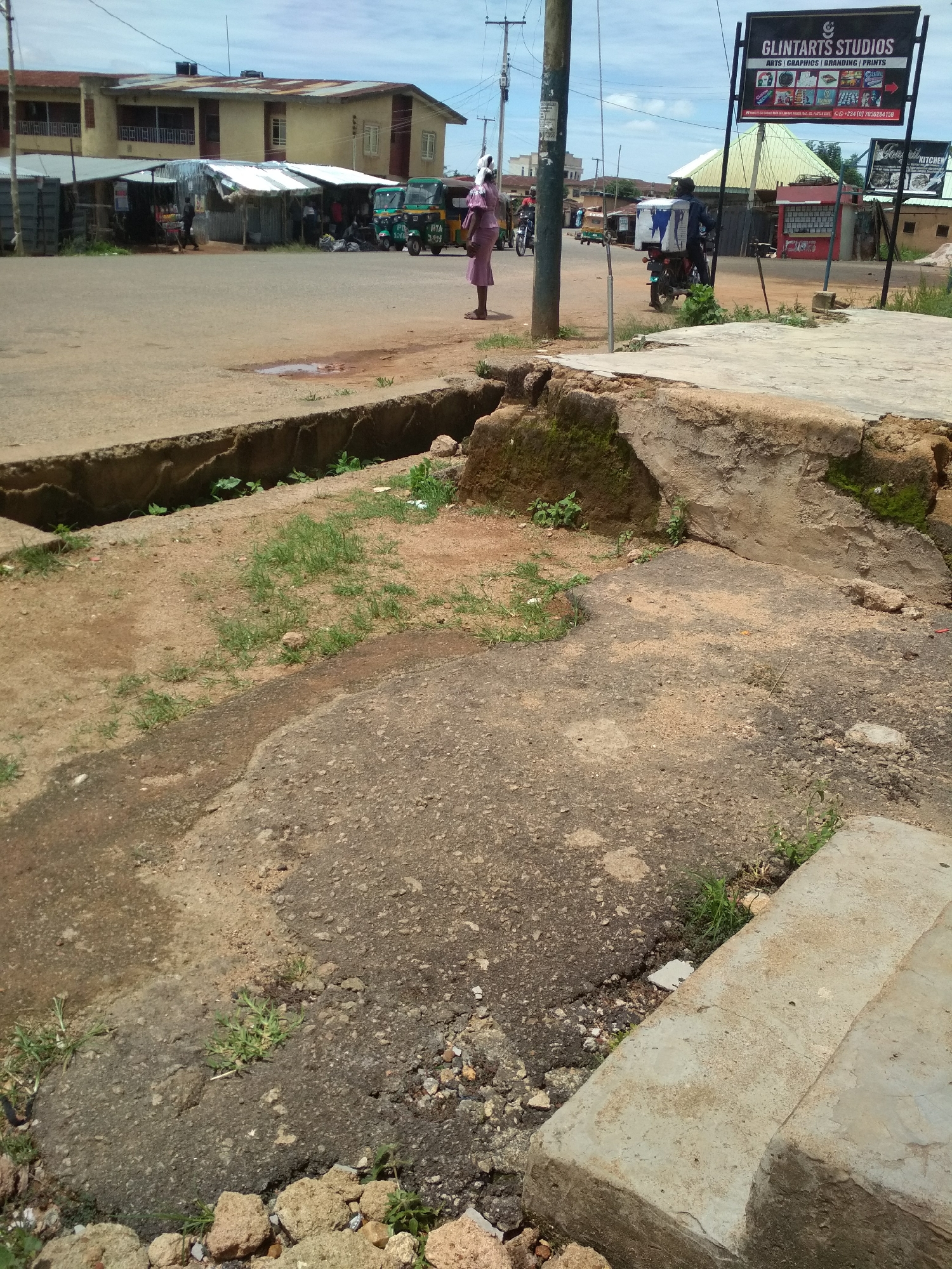 This detailed image captures an urban street scene in Jos, Nigeria, characterized by a mix of commercial activity and utilitarian infrastructure.
In the immediate foreground, a weathered concrete drainage channel runs horizontally, partially filled with dirt and sparse vegetation. To its right, the ground is a mix of packed dirt and deteriorated asphalt, featuring cracks, loose gravel, and small patches of grass. Large, irregular concrete blocks, possibly remnants of a curb or step, are visible at the bottom right.
The midground features a paved road, appearing worn with multiple patches, discolorations, and small potholes.
The left background is dominated by a two-story building with a light yellow or cream facade, multiple windows, and a reddish-brown corrugated metal roof. In front of this building, a row of smaller, single-story market stalls with corrugated metal roofs lines the roadside. Several people are visible near these stalls, engaged in various activities, and three green and white motorized tricycles (Keke Napep) are parked or positioned near the stalls. A person wearing a light pink or purple garment stands at the far right edge of this section, facing left.
The right background features a prominent black signboard for "GLINTARTS STUDIOS," advertising "ARTS | GRAPHICS | BRANDING | PRINTS" along with small icon representations of services and a Nigerian contact number (+234 (0) 7036264159). Below it, a smaller sign, partially obscured, shows text including "HOME KITCHEN." Behind these signs, a red kiosk-like structure is visible. A person on a motorcycle, carrying a large white delivery box on the back, is captured in motion, heading leftward past the signs. Overhead utility lines crisscross the sky, supported by visible utility poles. The sky is bright with some scattered clouds.