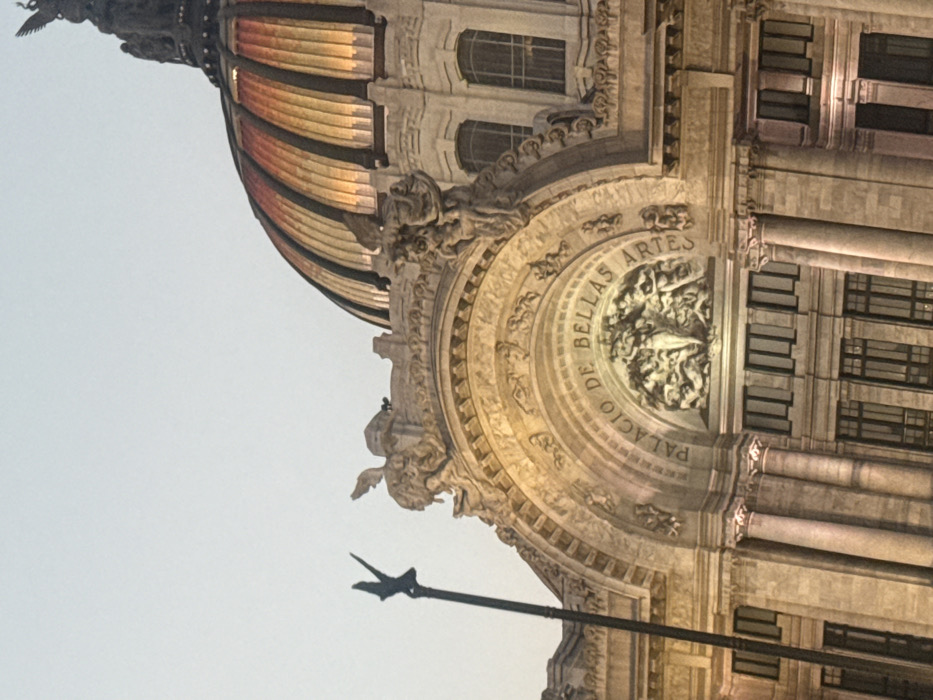 The image depicts a partial view of the Palacio de Bellas Artes. Ornate stone carvings adorn the building's facade, with the words "Palacio de Bellas Artes" inscribed in a semi-circular pattern above a sculpted relief. The building's dome exhibits radiating bands of color. Architectural elements like columns and windows are also visible, indicating a classical style. The angle suggests the photo was taken looking up towards the building.