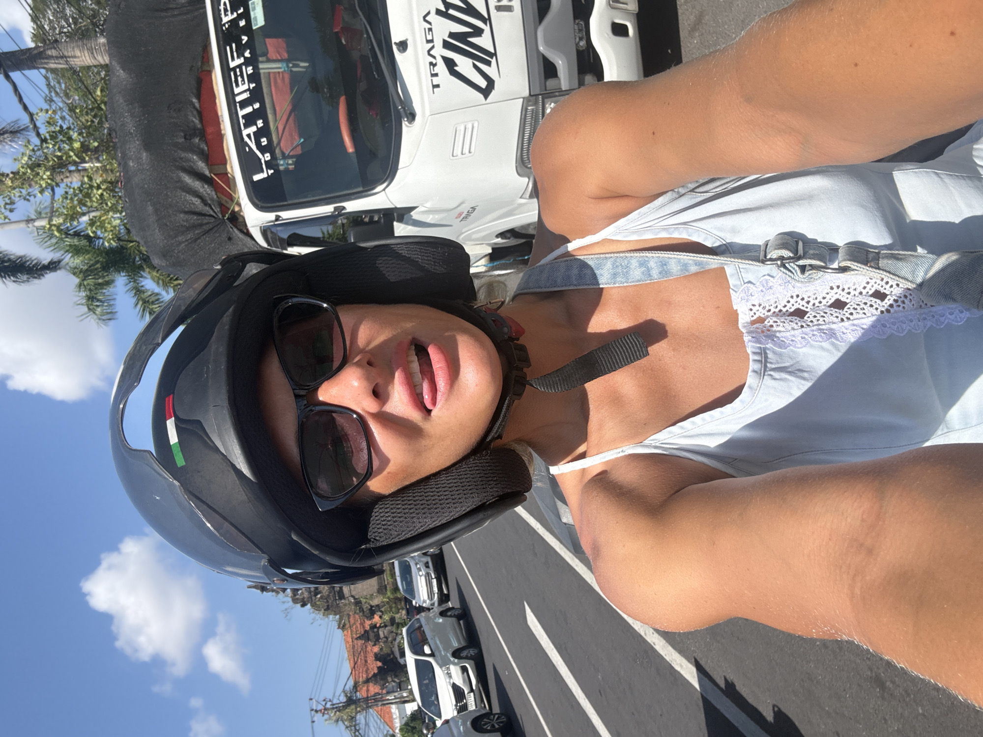 A woman wearing a helmet and sunglasses is taking a selfie while riding a scooter on a street. She is sticking her tongue out playfully. In the background, there are cars, a truck, and palm trees. The sky is blue with some clouds.
