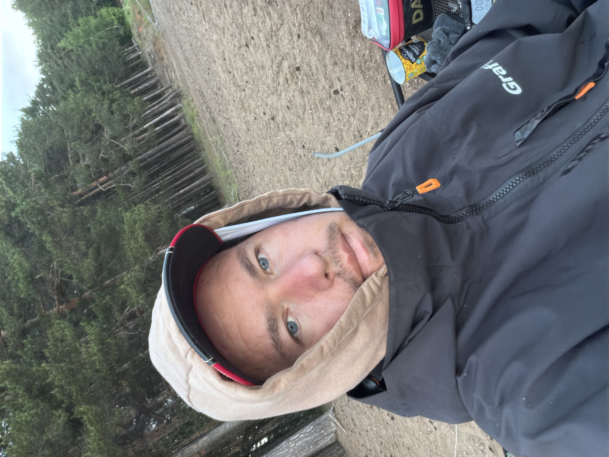 The image is a vertical, close-up selfie-style shot of a person outdoors in what appears to be a forest or wooded area, with the location context provided as Chaikovsky, Russia. The shot is taken from a slightly upward angle, showing the person's face prominently.
The primary subject is a man with blue eyes, light skin, and stubble on his chin. He is looking directly at the camera with a neutral expression. He is wearing a black jacket with the hood pulled up, and under the hood, a light brown or beige hoodie is visible. A baseball cap with a black brim and red trim is also visible under the hoodie.
In the background, to the left of the person, is a dense forest of evergreen trees, with many fallen logs on the ground, suggesting a natural, possibly rugged, environment. To the right and below the person, the ground is visible, appearing somewhat muddy or earthy.
Several objects are visible around the person. On the right side of the image, next to the person, parts of what looks like camping or outdoor gear are present. A black jacket, possibly the same one the person is wearing, has a white logo that reads "Graff" on the sleeve. Orange zipper pulls are also visible on the jacket. Near the jacket, a small red and white pouch is partially seen, with the letters "DAI" visible on it. Below this, a silver can with a yellow label is present, on which the word "GRENOLIA" and "RYŻOWY" are legible. A gray cable or tube runs across the ground nearby.
The overall impression is of someone taking a break in a natural, outdoor setting, possibly during a cool or overcast day, given the warm clothing.