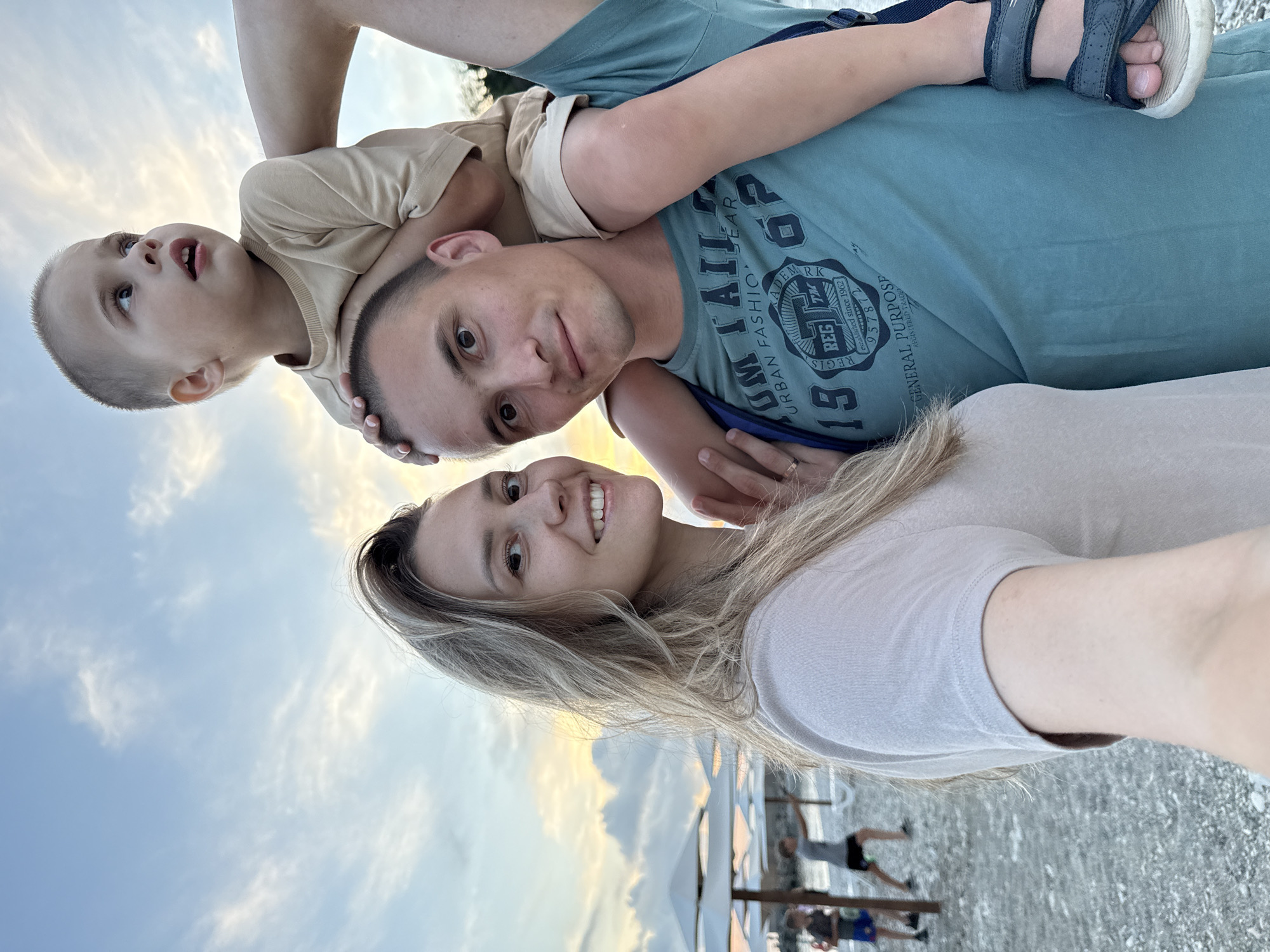 A family photo taken on the beach in Nebug, Russia, shows a young boy sitting on his father's shoulders. The father, wearing a teal t-shirt with text graphics, looks directly at the camera. The mother, to the left, also smiles at the camera. They are on a pebble beach with umbrellas and other beachgoers in the background. The sky is cloudy with a sunset glow.
